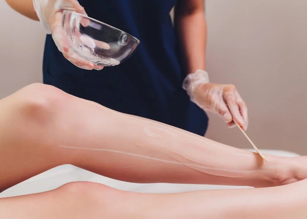 Technician spreading wax on a client’s lower leg with a wooden stick while holding a glass bowl, demonstrating pre-wax care.