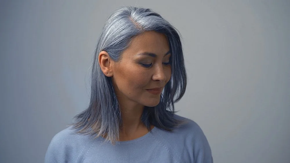 A woman with smooth silver-gray hair, looking down softly indoors, showcases the best hair color for gray hair.
