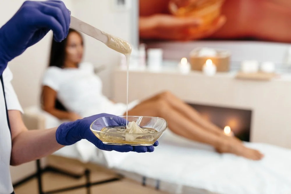 An esthetician wearing blue gloves holds a bowl of warm wax and a wooden applicator while a client lies on a spa treatment bed in the background.
