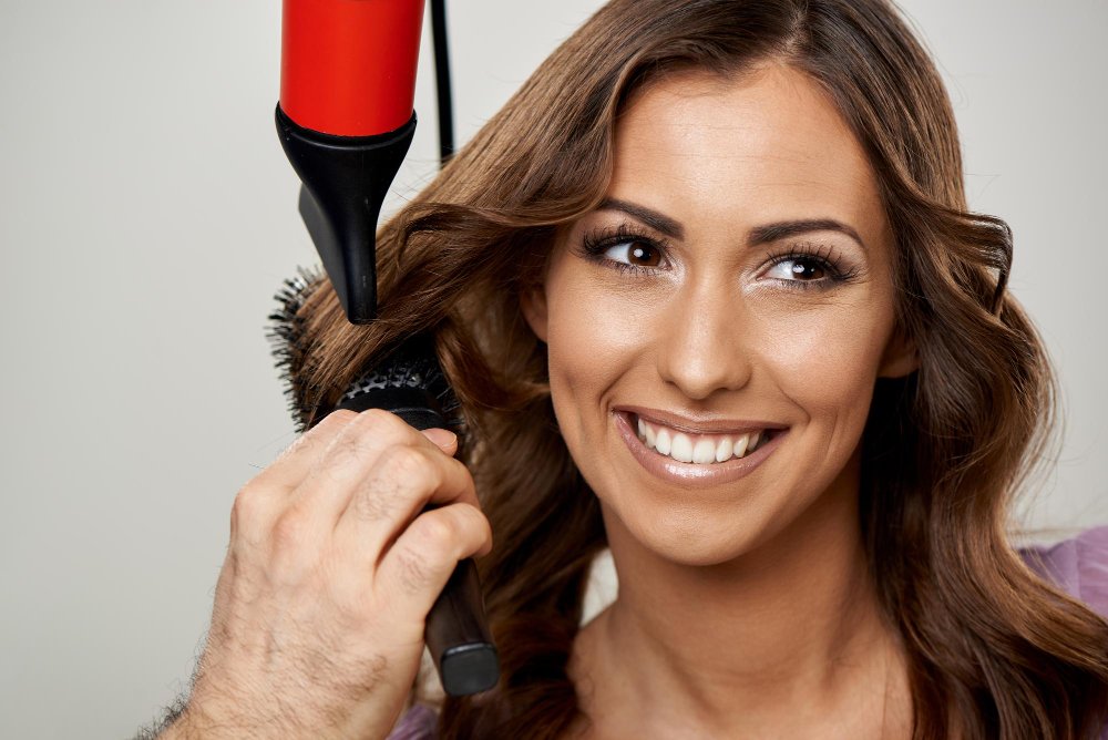 A stylist using a round brush and a hair dryer to lift and shape hair for added volume.