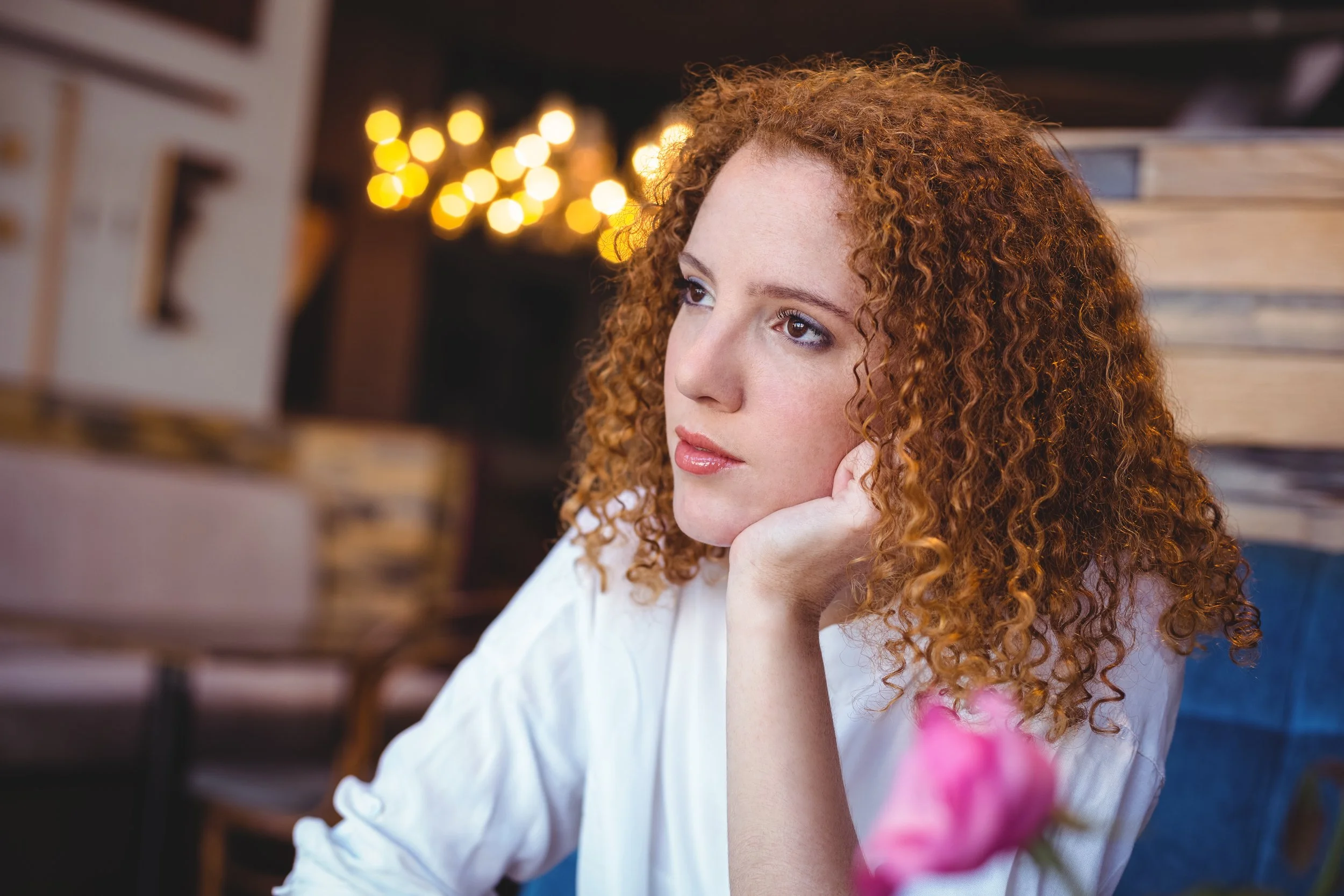 A woman with curly red hair resting her chin on her hand at a cafe table.