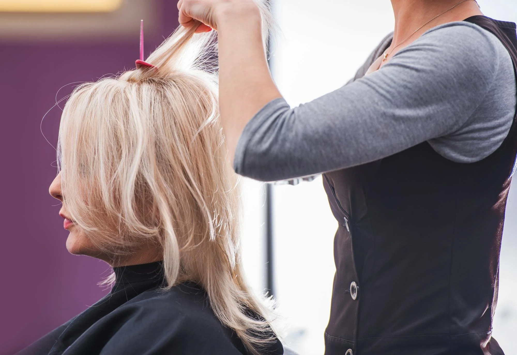 A stylist carefully sectioning a blonde client's hair with a pintail comb for ammonia-free professional hair color.