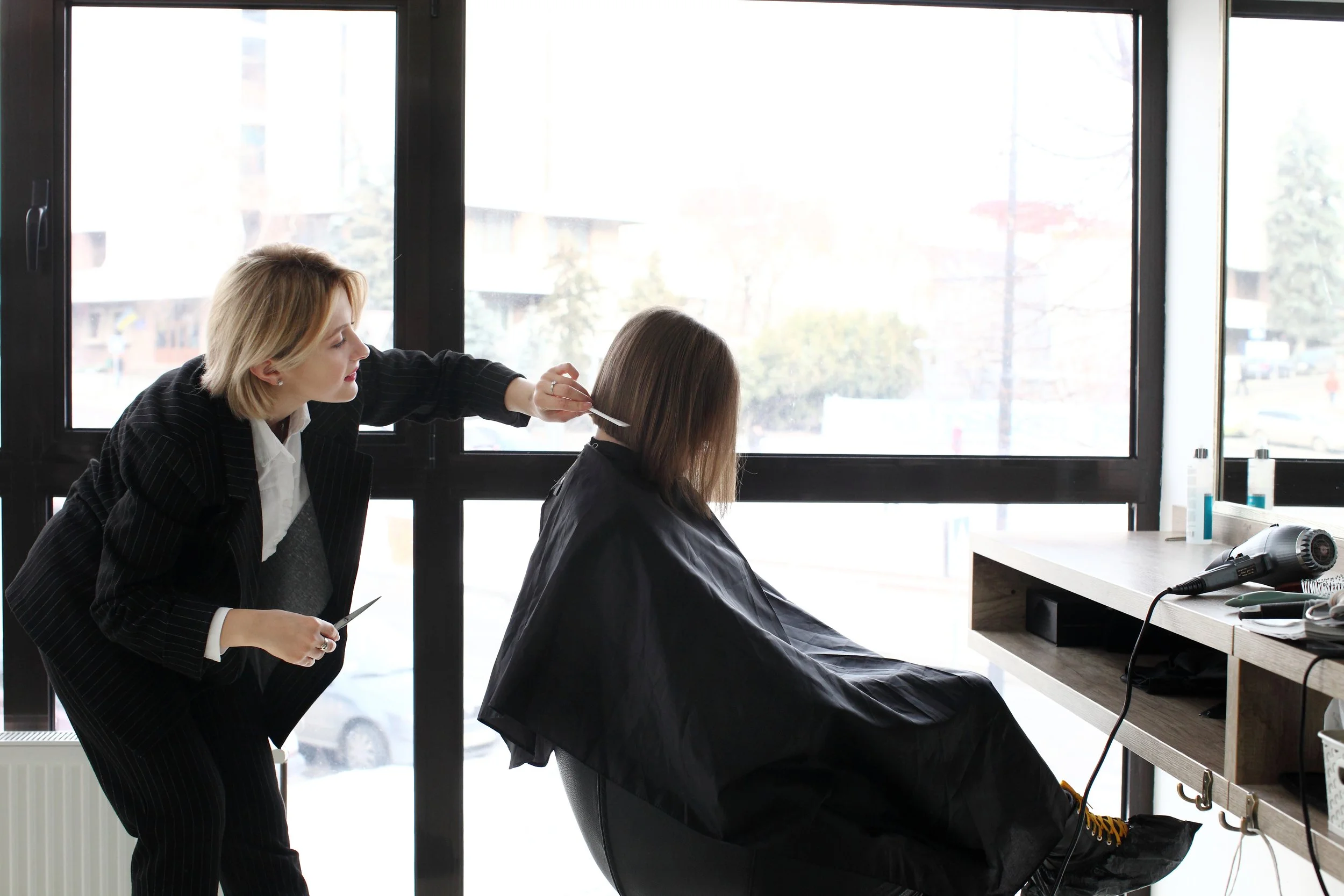 A stylist cutting a client's hair near a bright window in a modern salon.