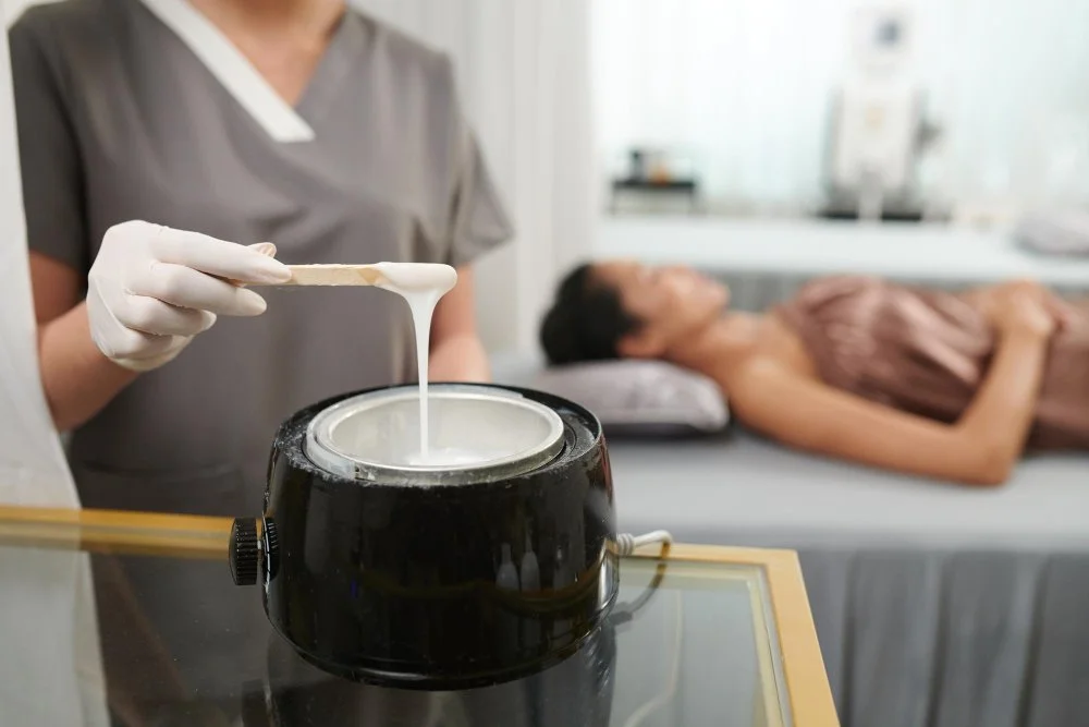 An esthetician dips a wooden applicator into a professional wax warmer while a client lies on a treatment table in a spa room.