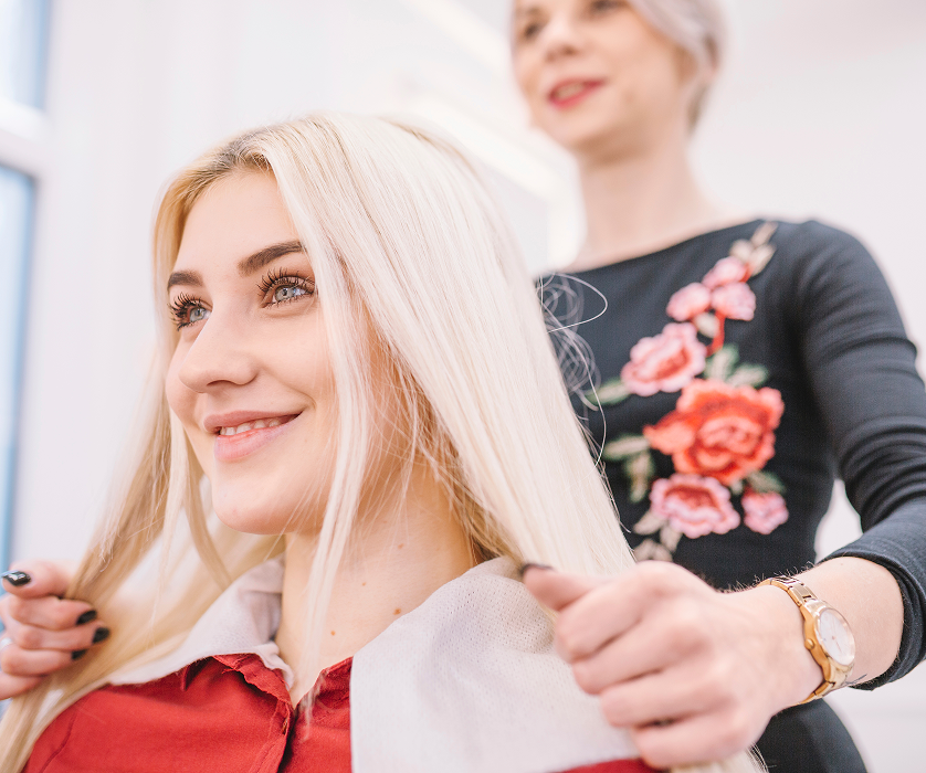 A smiling client seated in a salon chair while a hair stylist works on her blonde hair.