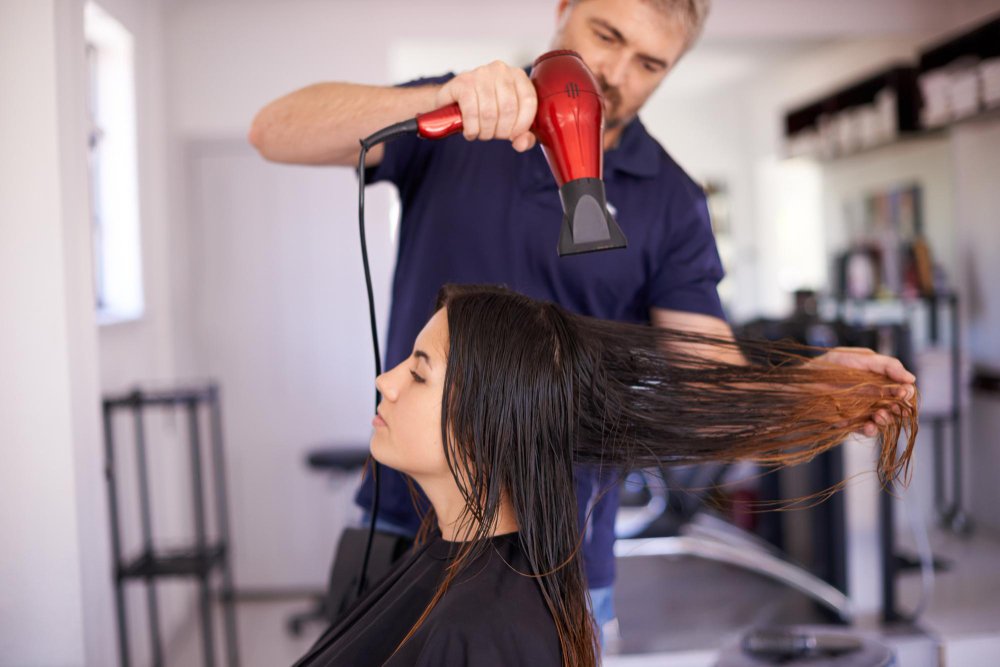 A stylist blow-drying long hair in sections using a brush and dryer.
