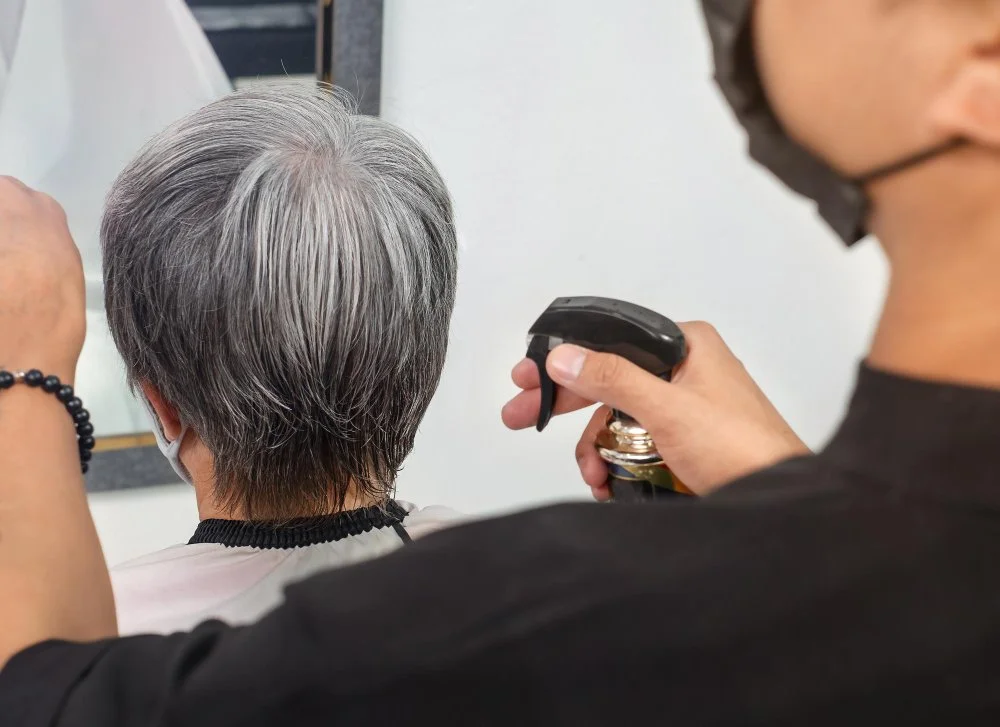 A stylist sprays product on short gray hair during a salon appointment, demonstrating how to color gray hair.