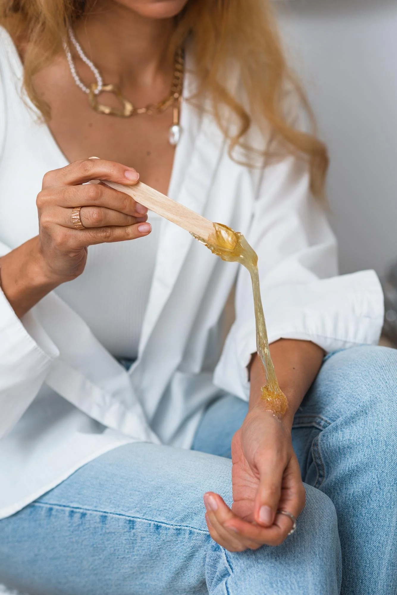 A woman in a white shirt holding a wooden spatula with dripping sugar wax, demonstrating sugar wax vs regular waxing.