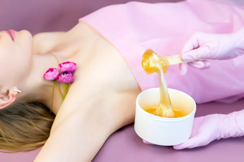 A client lies on a treatment table while an esthetician applies warm wax from a bowl using a wooden applicator.