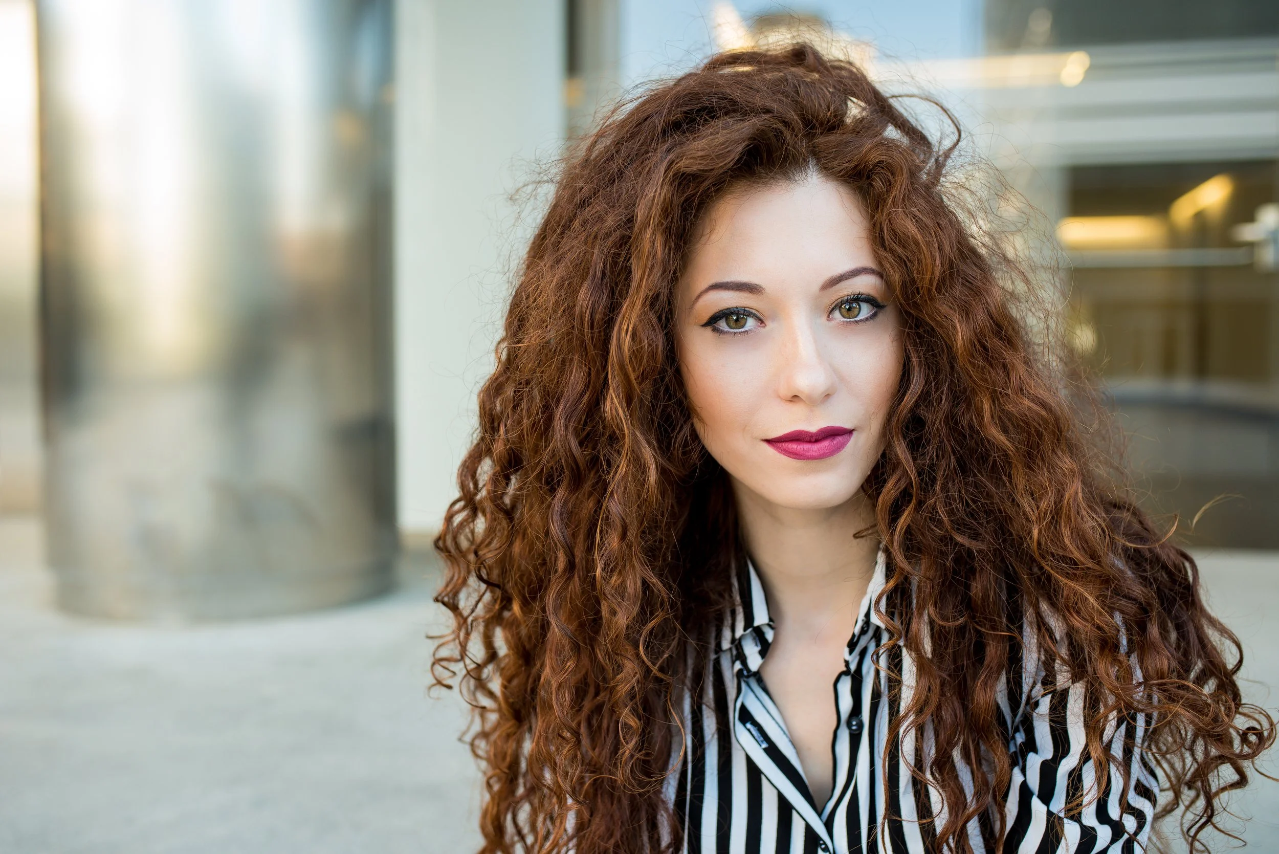A woman with long curly brown hair wearing a striped blouse outdoors.