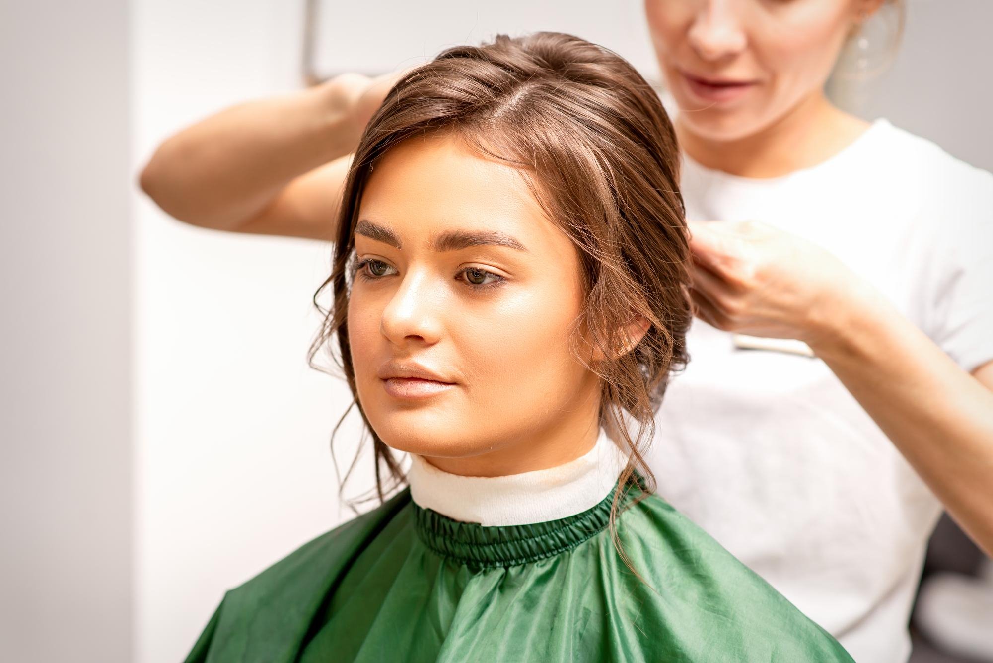 Young woman sitting in a salon chair with a green cape, having her hair styled into an elegant updo by a female hairdresser in a beauty salon.