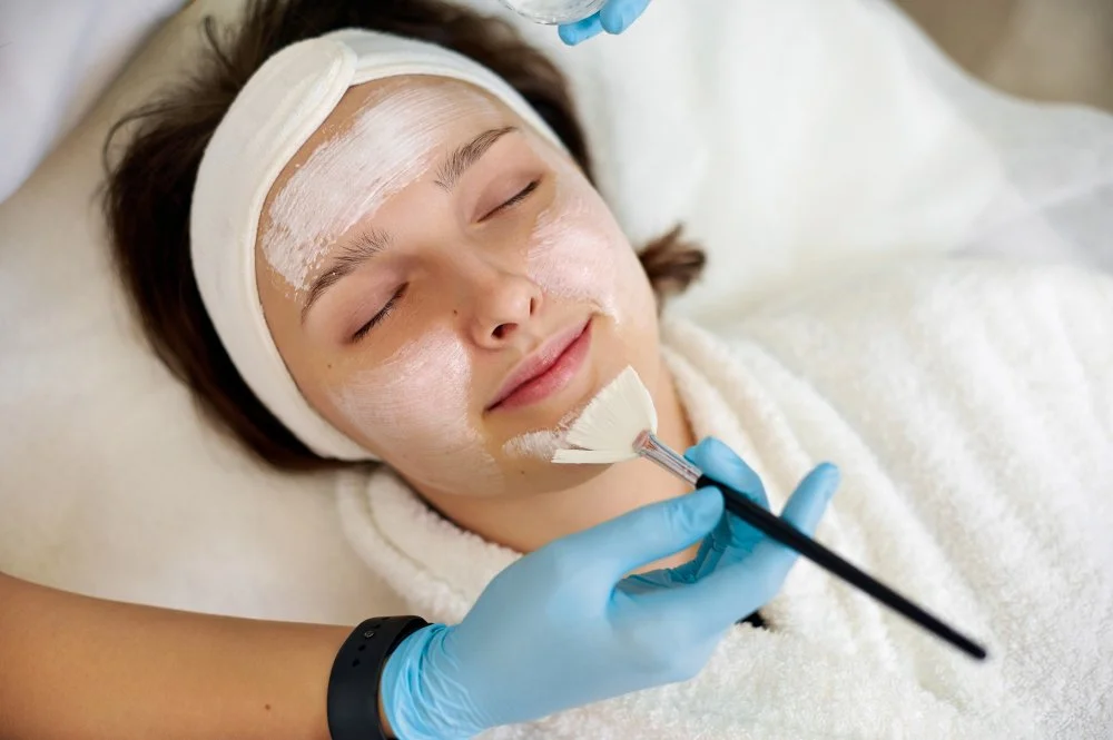 Gloved esthetician brushing treatment mask on a relaxed client’s face.