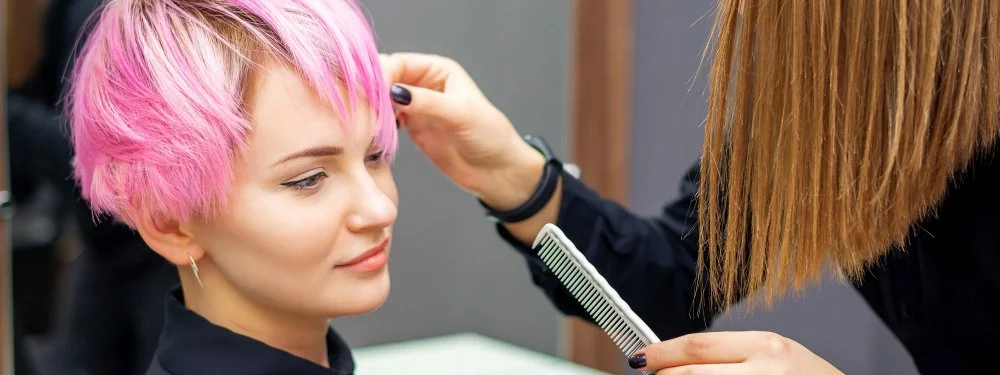Hairstylist adjusting and trimming short pink hair with a comb while the client sits in a salon chair.