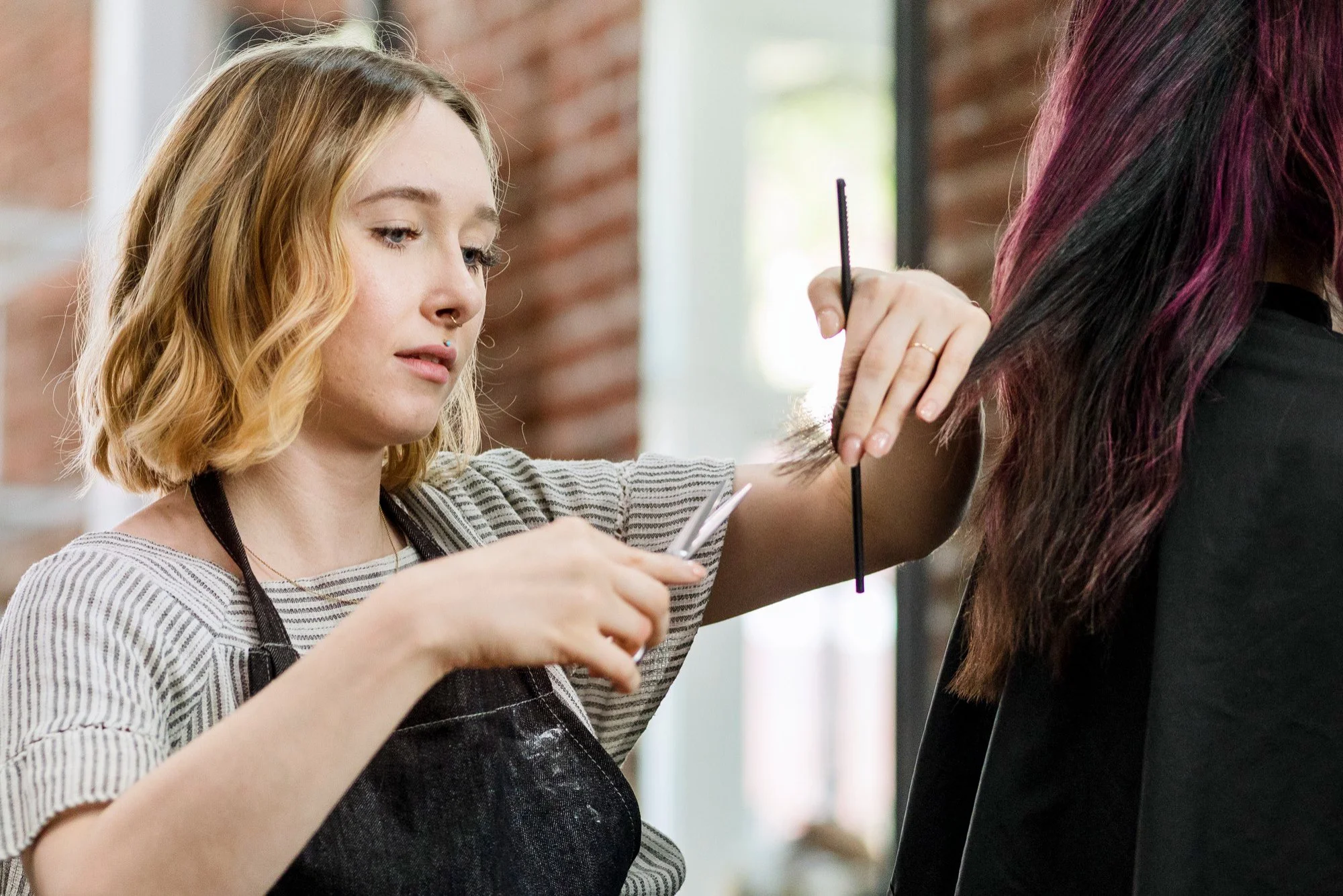 A customer sits in a beauty salon chair as a hairstylist trims her hair with scissors, illustrating the process of a professional woman’s haircut.