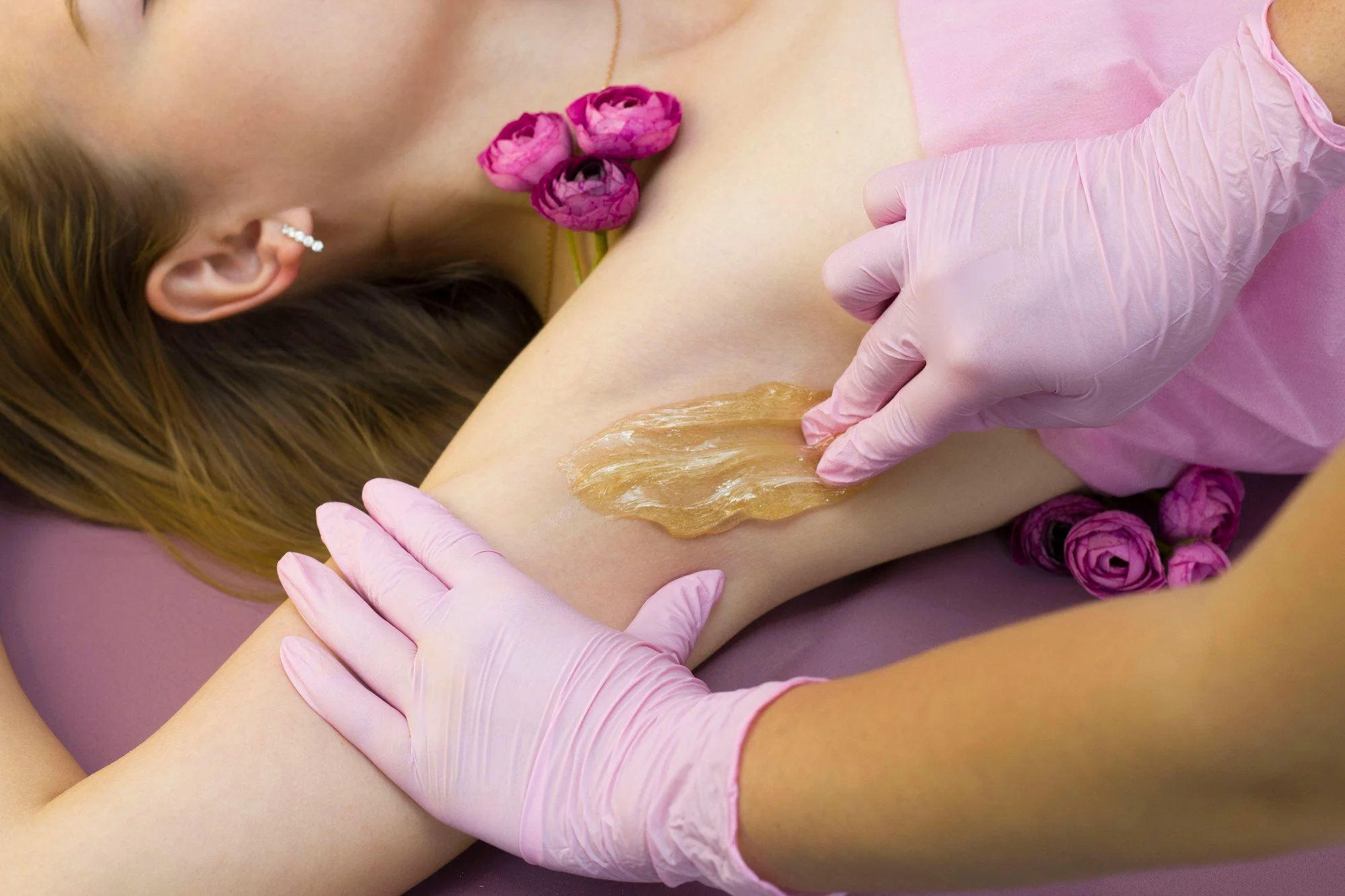 Esthetician in pink gloves applying warm wax to a client's underarm area for Brazilian wax aftercare.