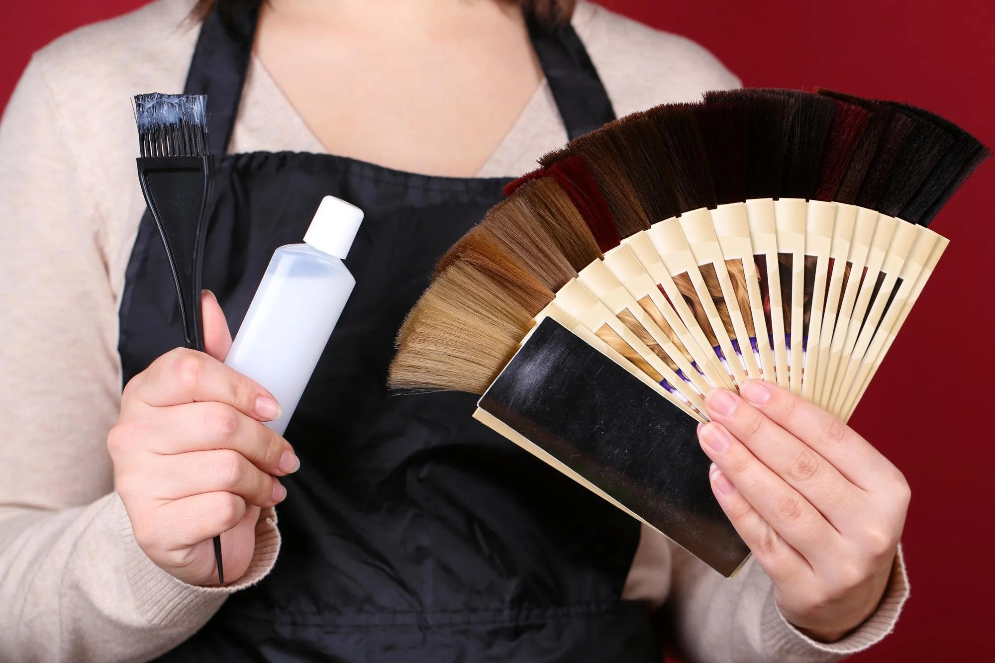 Colorist in a black apron holding a dye brush, a developer bottle, and a fan of swatches for a professional hair color service.