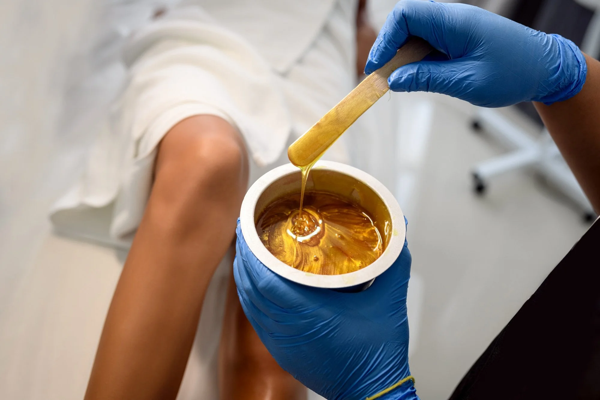 An esthetician in blue gloves is stirring warm wax with a wooden spatula next to a client's leg.