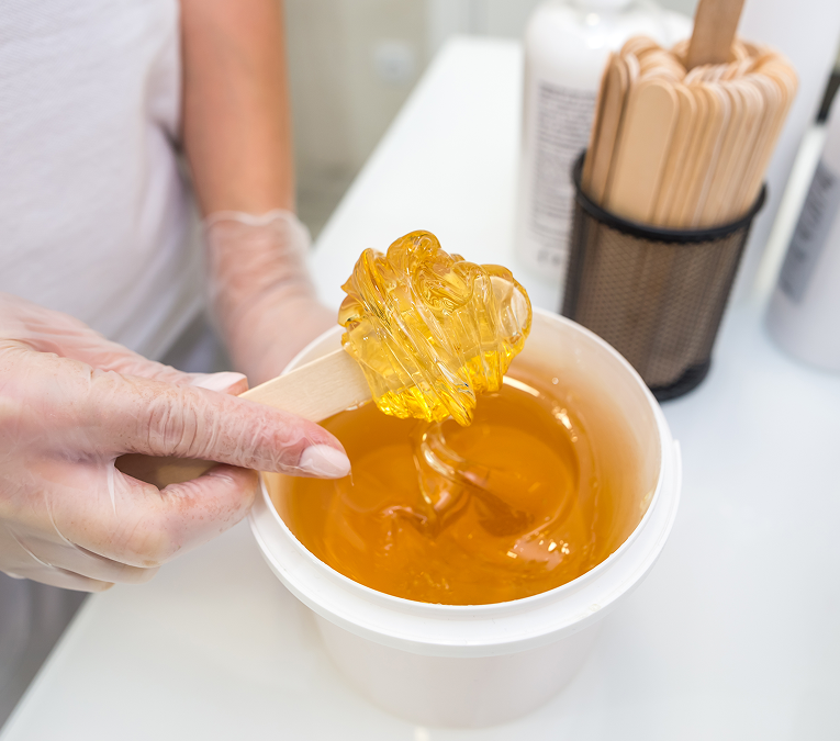 Sugar wax Chicago esthetician dipping a wooden spatula into warm golden sugar wax beside a client.