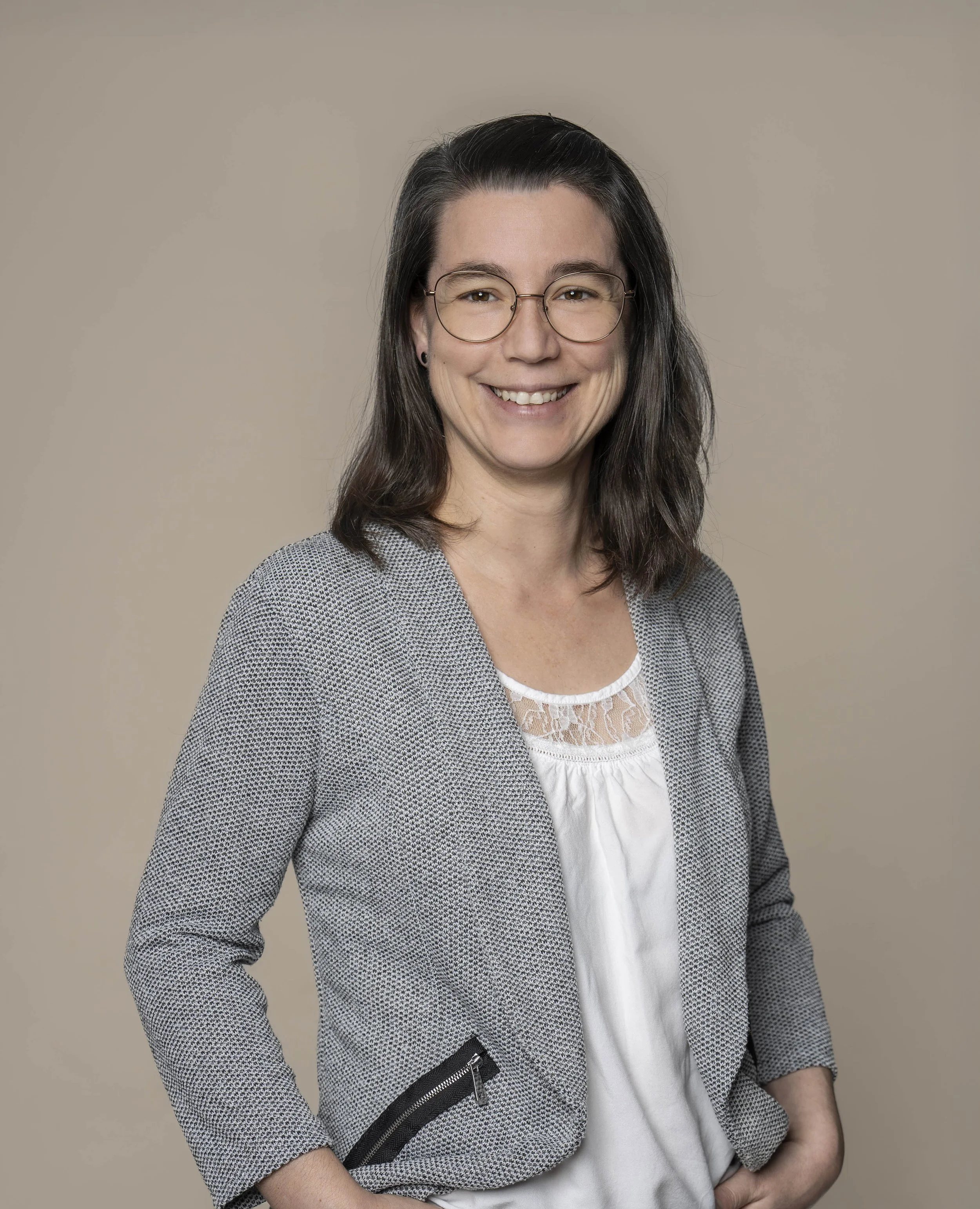 Porträt einer lächelnden Frau mit Brille, dunklen Haaren und einem grauen Blazer vor beige Hintergrund.