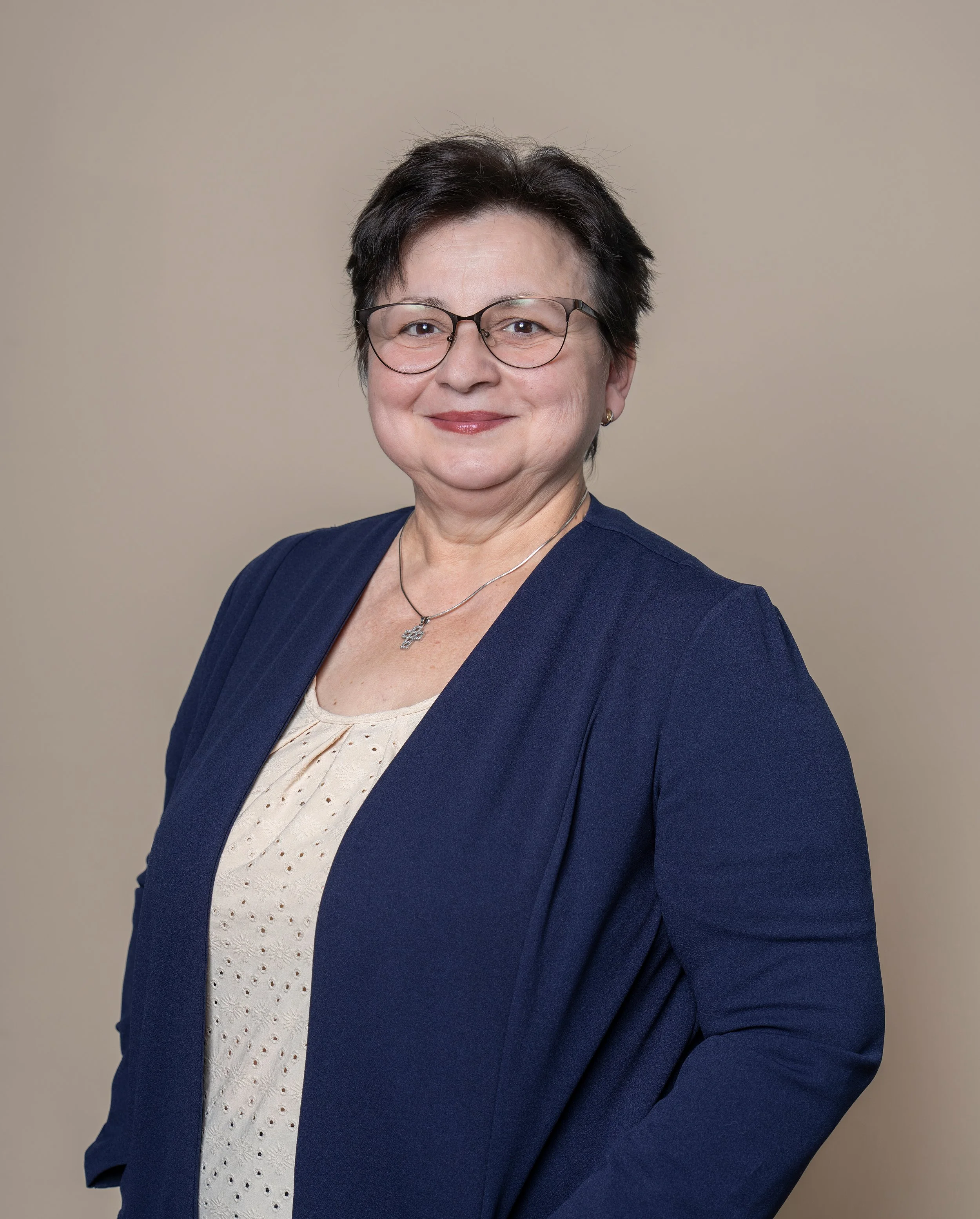 Eine Frau mit kurzen dunklen Haaren, Brille und einem dunkelblauen Blazer steht vor einem hellbeigen Hintergrund.