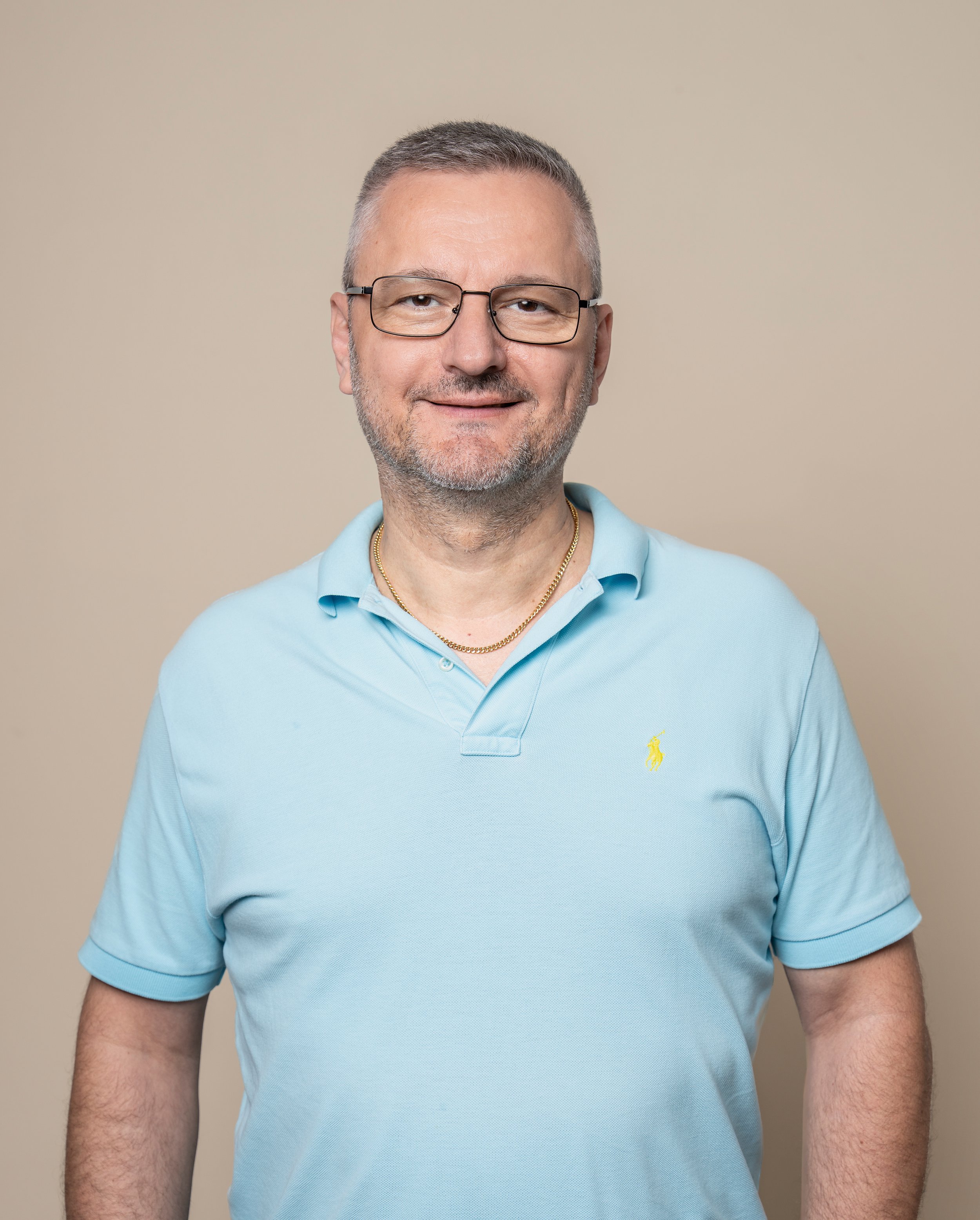 Mann mit Brille, kurzen grauen Haaren, grauem Bart, in hellblauem Polo-Shirt, stehen vor beige Hintergrund.