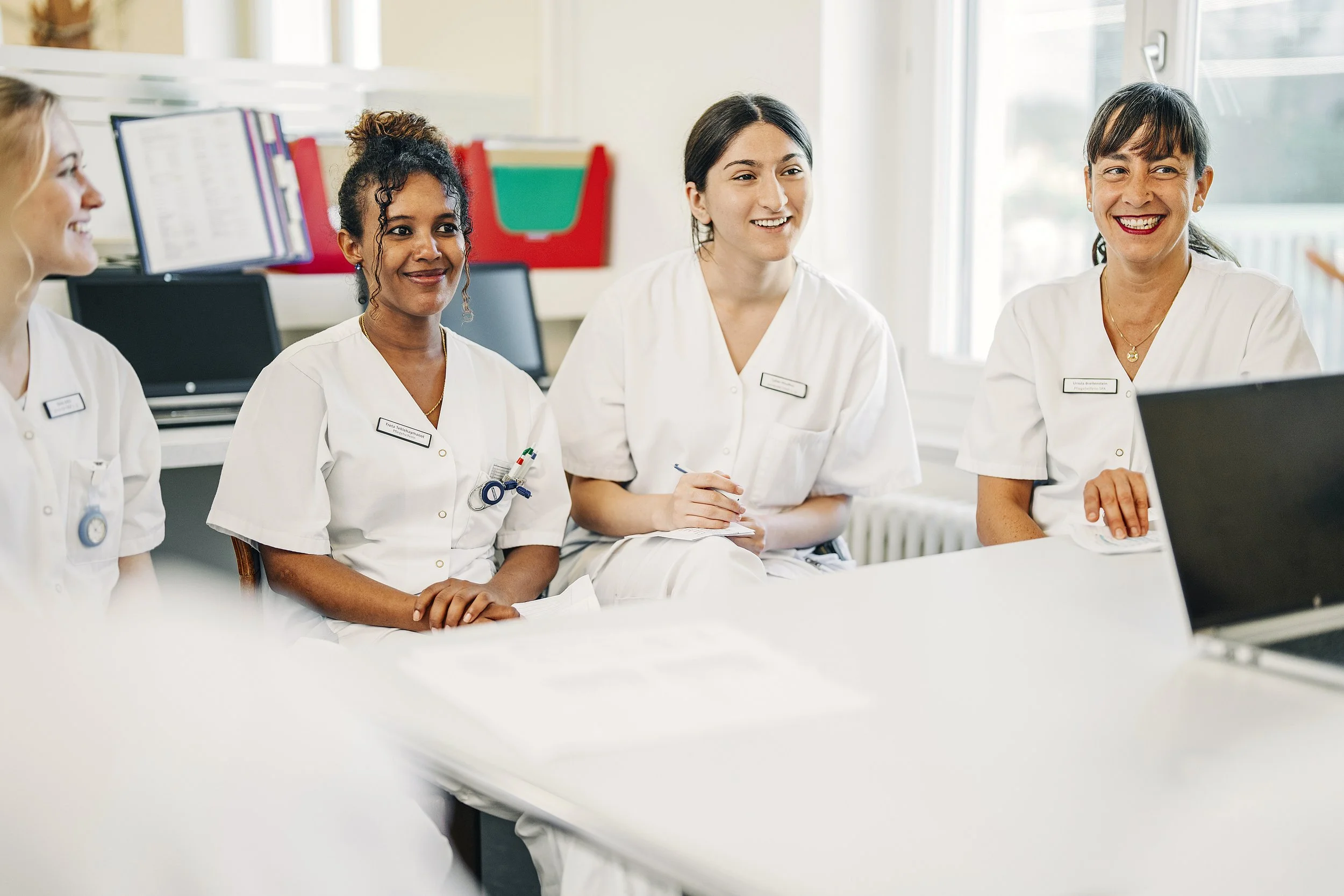 Vier Frauen in weißen medizinischen Kitteln vom Pflegepersonal sitzen in einem Besprechungsraum und lachen.