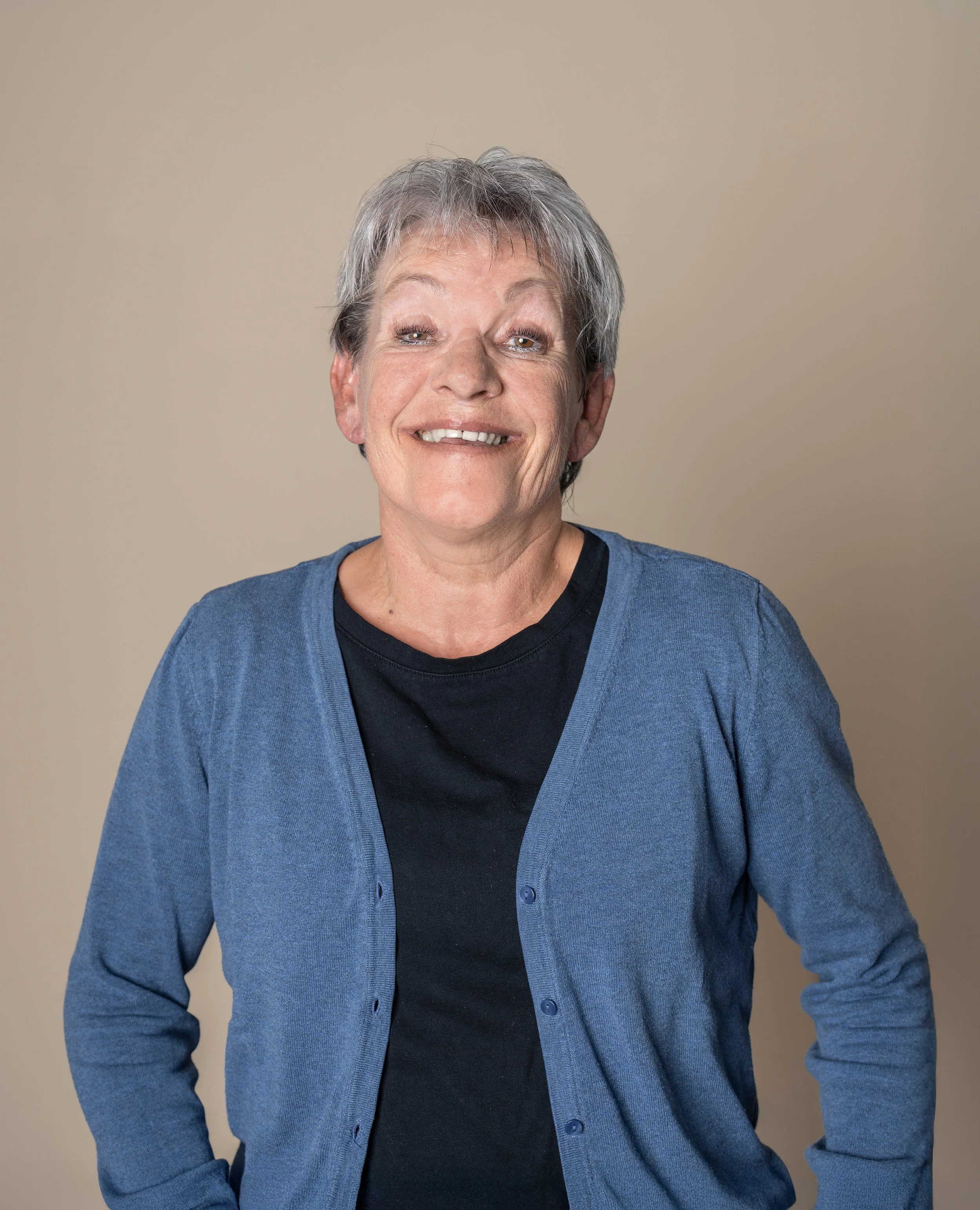 Eine ältere Frau mit kurzen grauen Haaren, lachend, vor einem neutralen Hintergrund, trägt eine schwarze Bluse und eine blaue Strickjacke.