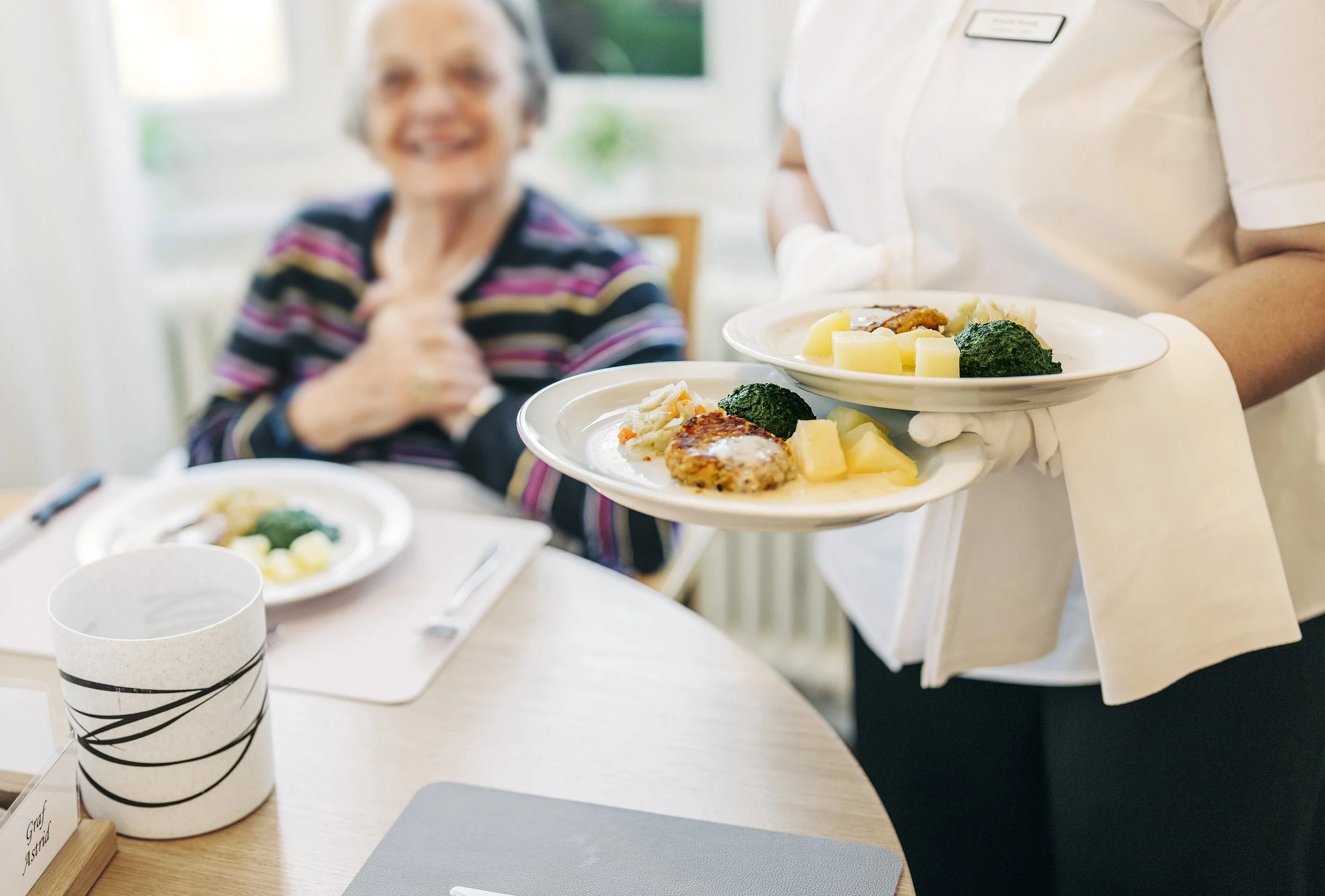 Eine pflegekräfte serviert Essen auf Tellern vor einer lachenden älteren Frau im Pflegeheim.