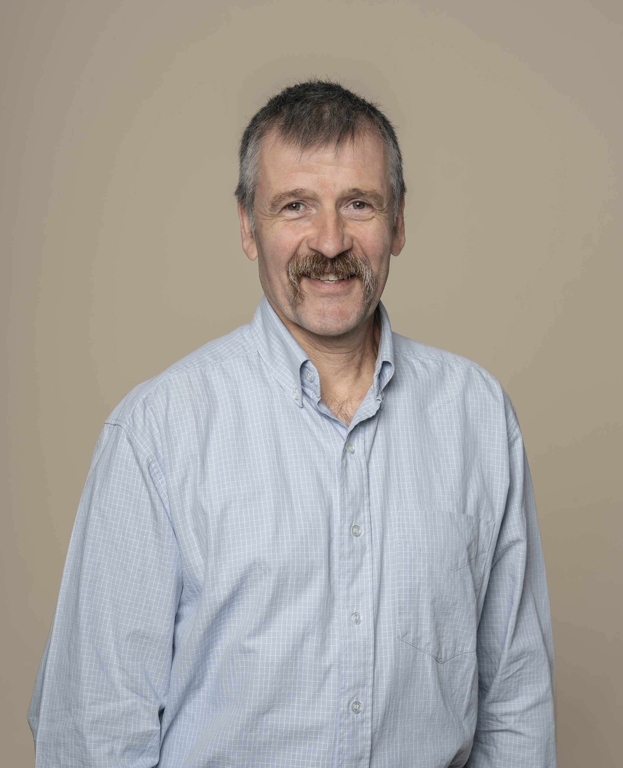 Ein Mann mit grauem Haar, Kinnbart und Tanker, trägt ein blau-weiß gestreiftes Hemd, lächelt vor neutralem Hintergrund.