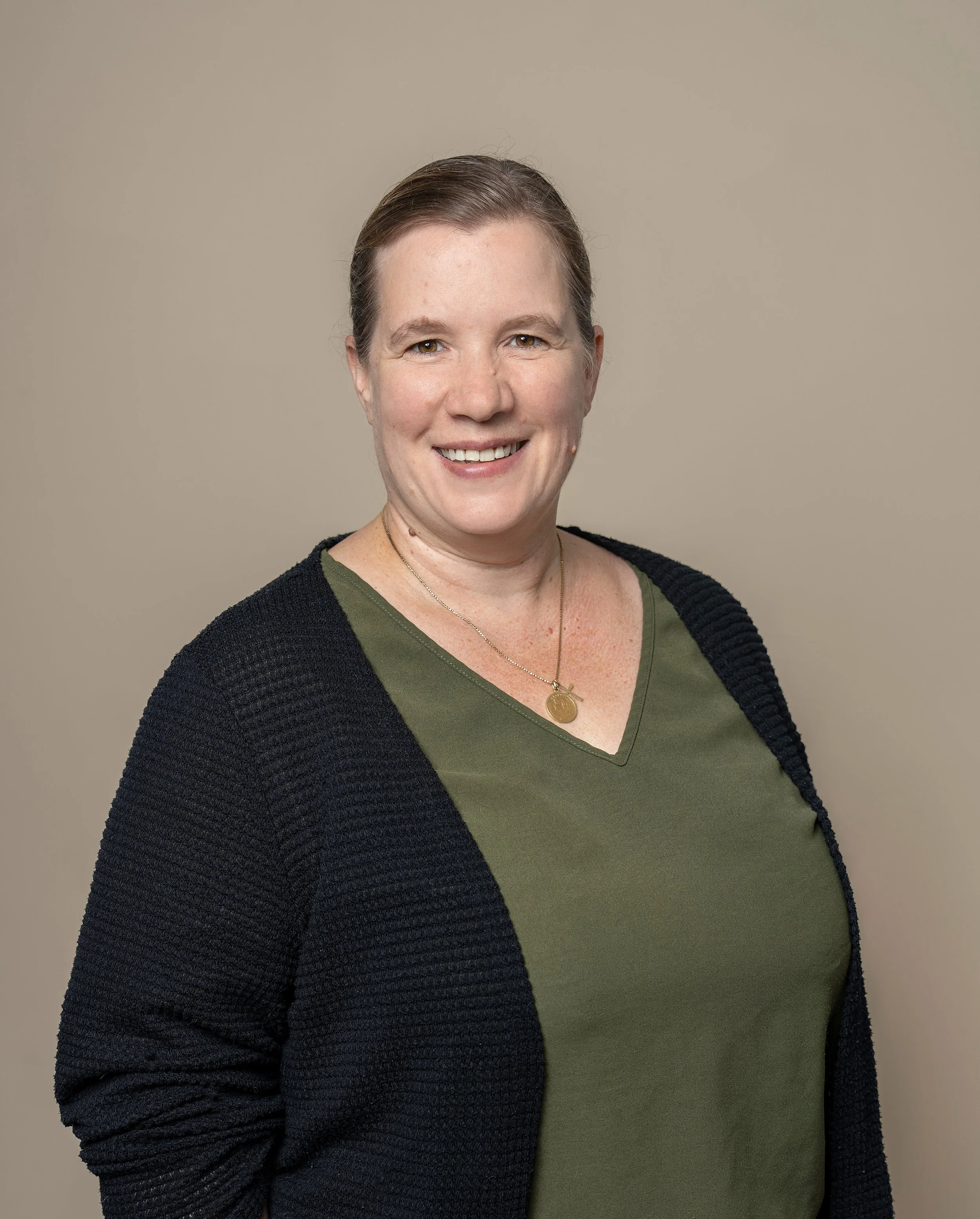 Porträt eines lächelnden Frau mit braunem Haar, grüner V-Ausschnitt-Shirt, schwarzer Strickjacke und goldener Halskette vor neutralem Hintergrund.