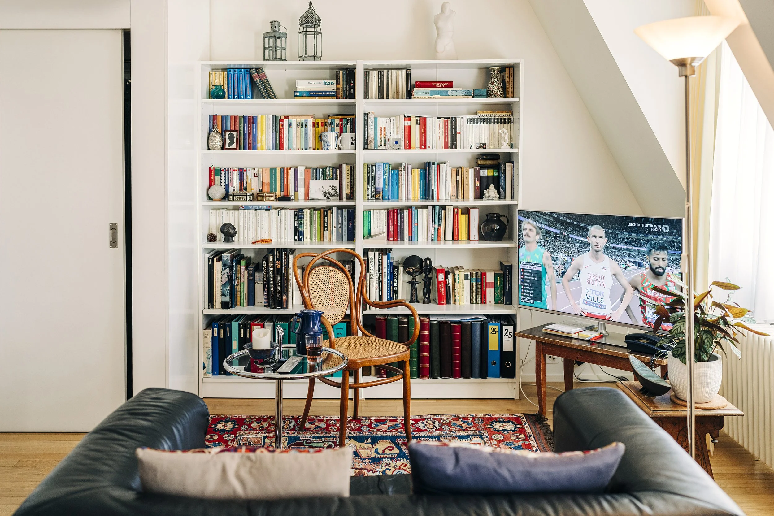 Gemütliches Wohnzimmer mit großer Bücherwand, Sessel, Couchtisch mit Kerzen, Fernseher, Stehlampe, Couchtische, Sofa mit Kissen, Teppich und Pflanze.