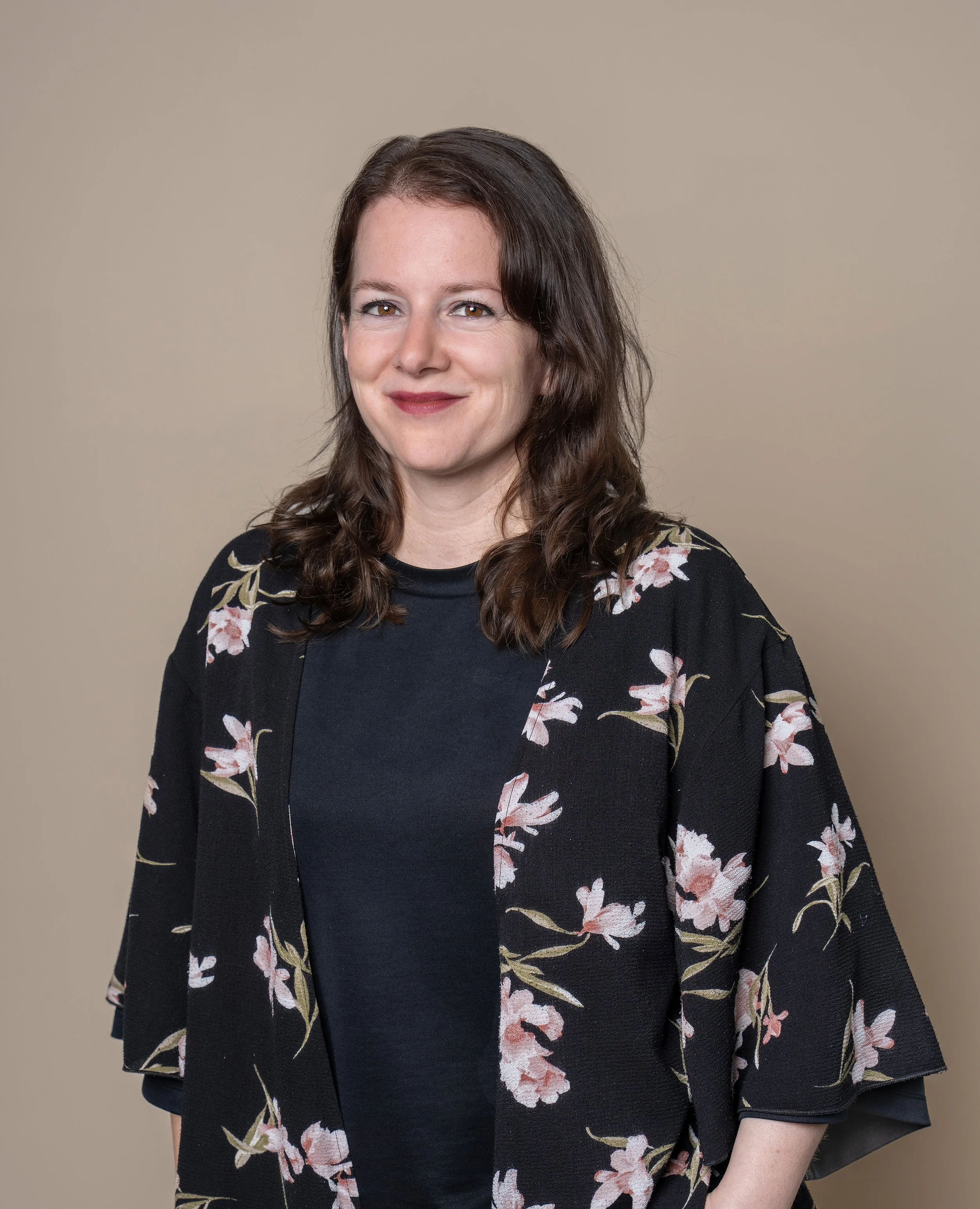 Porträt einer lächelnden Frau mit schulterlangem braunem Haar, schwarzem Top und blumigem Kimono vor beigem Hintergrund.