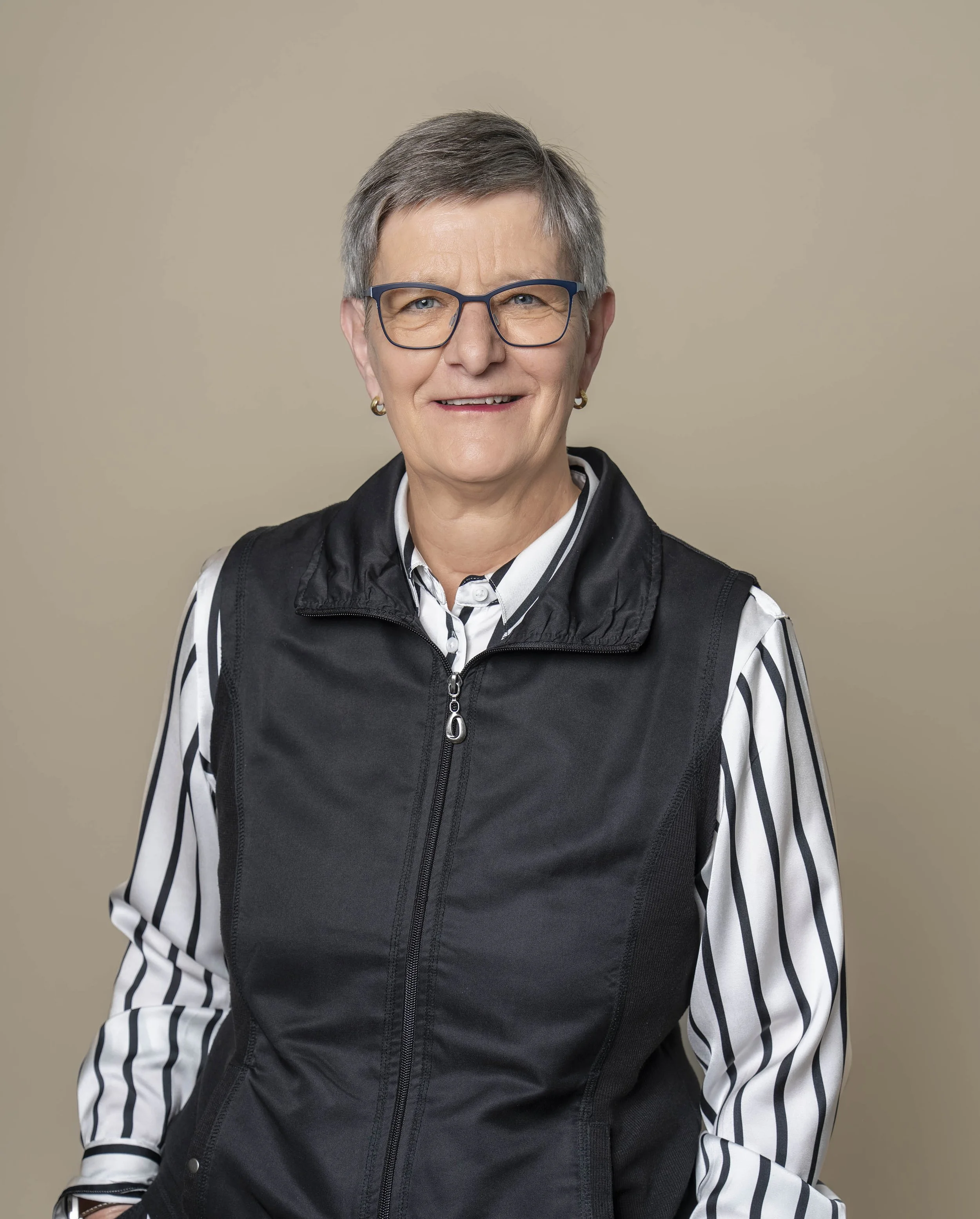 Porträt einer älteren Frau mit Kurzhaarschnitt, Brille, schwarzer Weste und gestreiftem Hemd vor neutralem Hintergrund.