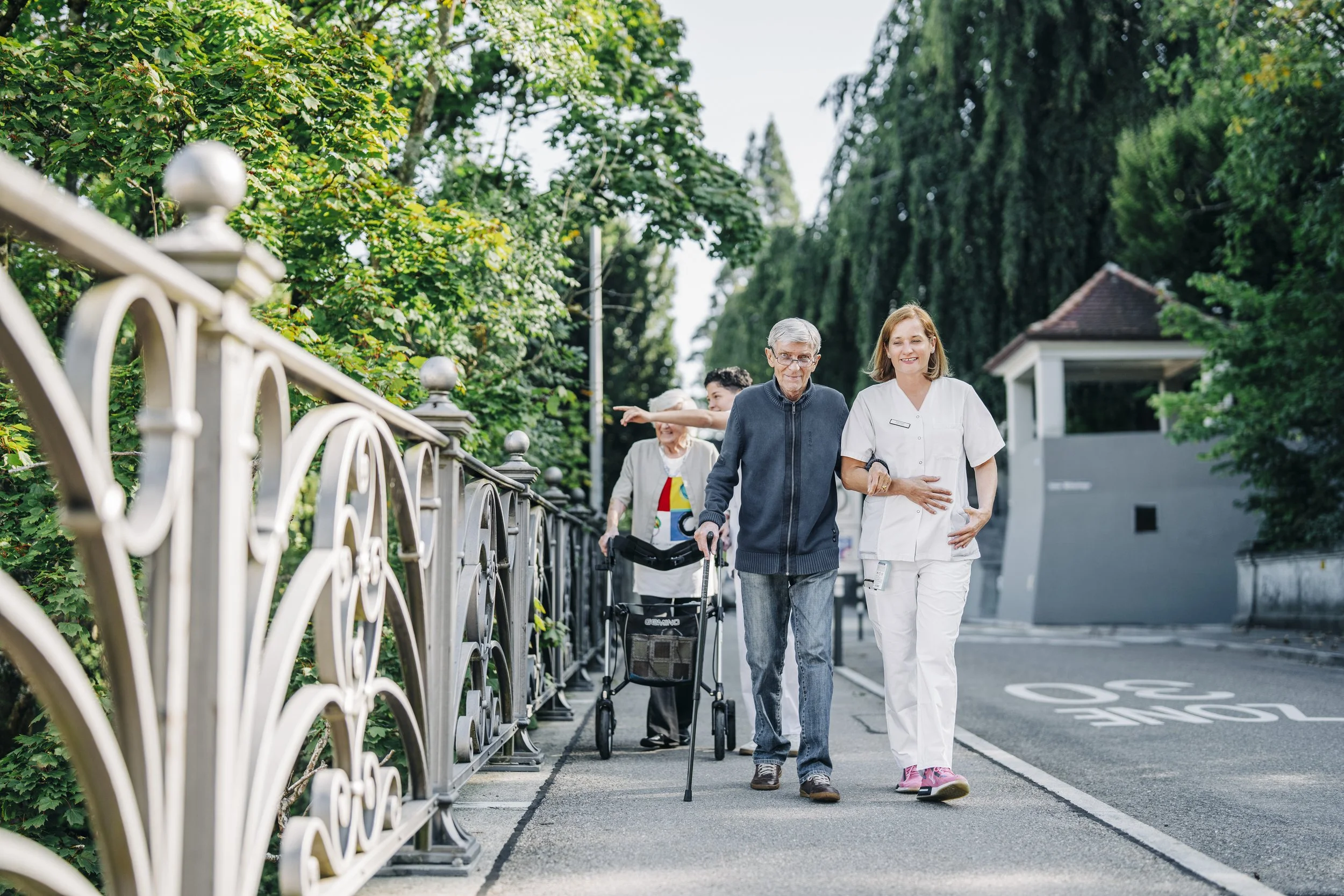 Pflegepersonal begleitet älteren Menschen auf dem Spaziergang, im Hintergrund weitere Personen und eine grüne, bewaldete Umgebung.