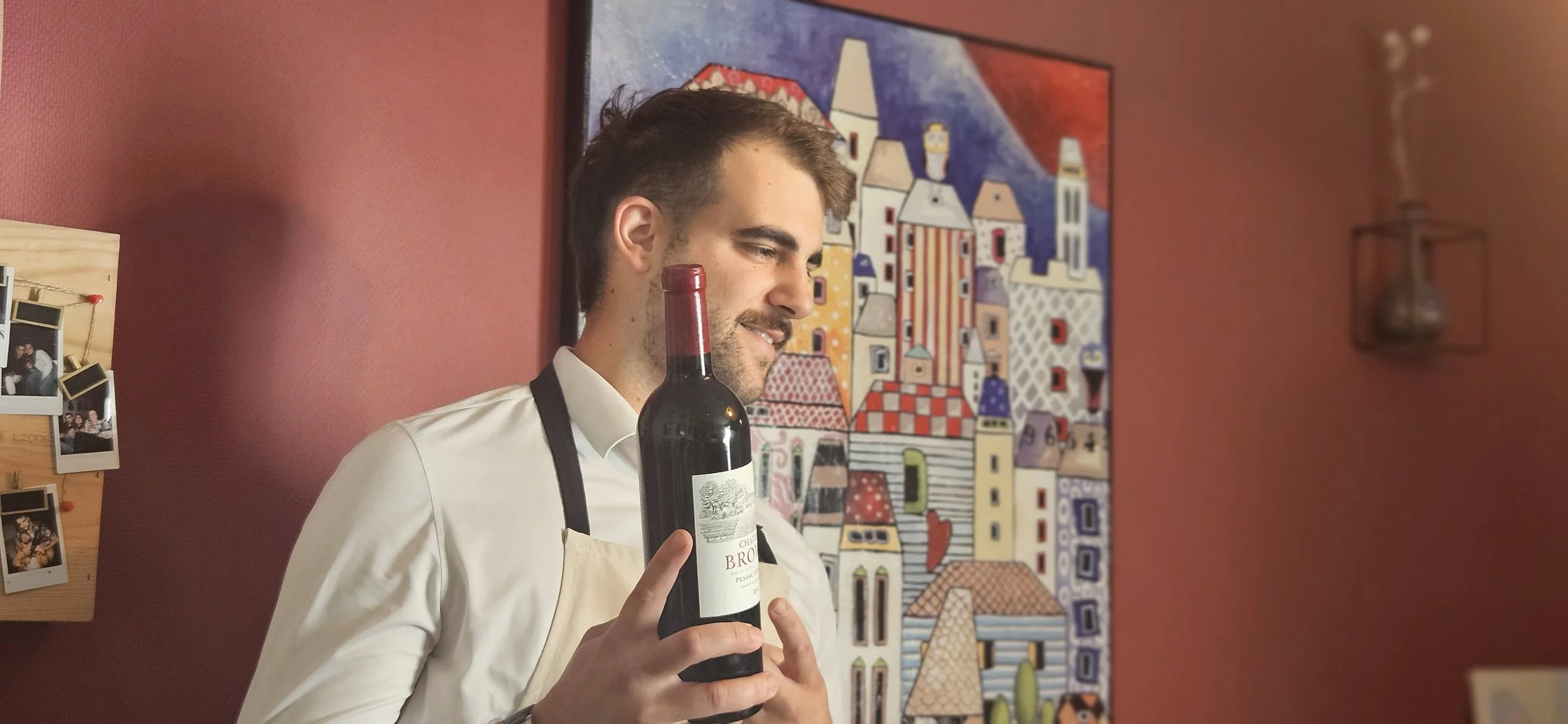Un jeune homme souriant tenant une bouteille de vin rouge, habillé d'une chemise blanche et d'un tablier beige, dans une pièce avec un mur rouge et une peinture colorée en arrière-plan.