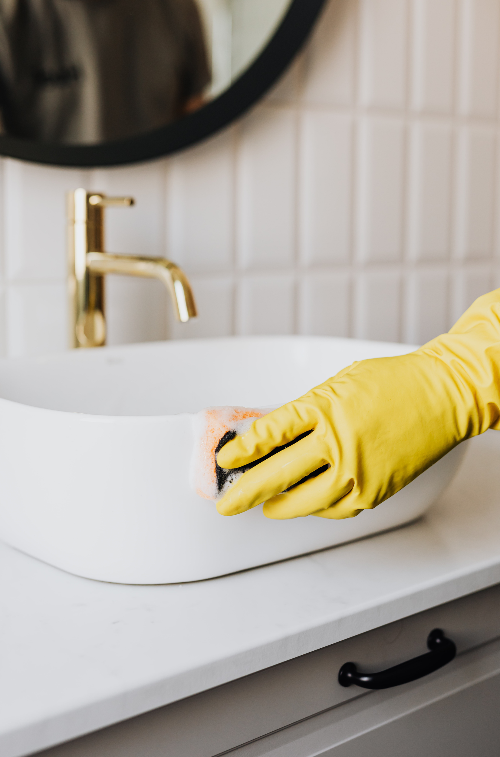 Person wearing yellow rubber gloves cleaning a white ceramic sink with a yellow sponge in a modern bathroom.