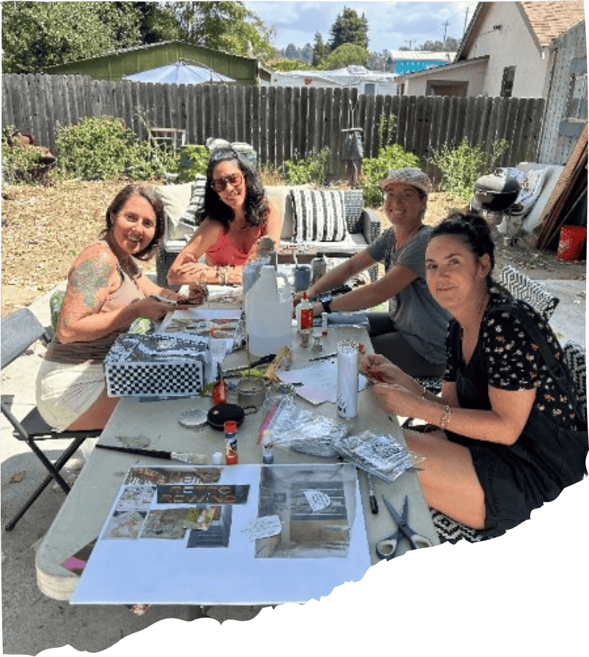 Four women sitting at a table outdoors, celebrating with food and drinks in a backyard with a wooden fence, plants, and patio furniture.
