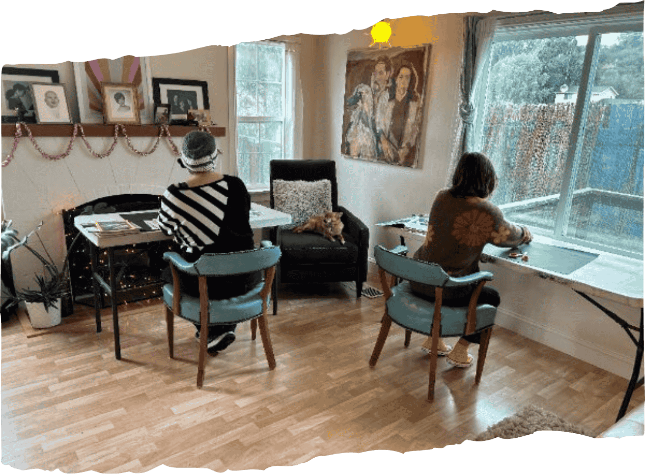 Living room with two women sitting at different tables, one working on a laptop, the other looking out the window, with a black armchair, family photos, and artwork on the walls, during daytime.