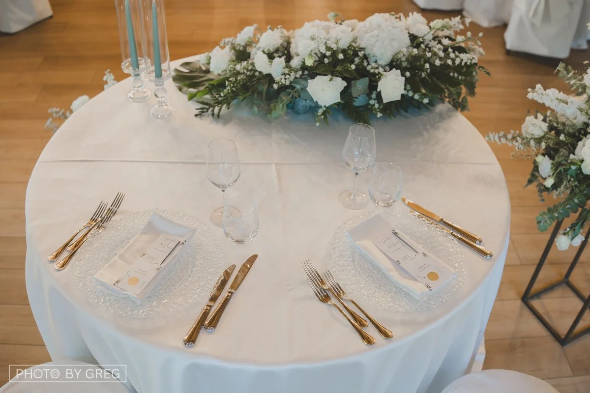 Tavola di matrimonio nei toni del bianco e dell’oro, con fiocchi sui tovaglioli e composizione floreale. Gratta e vinci al tovagliolo.