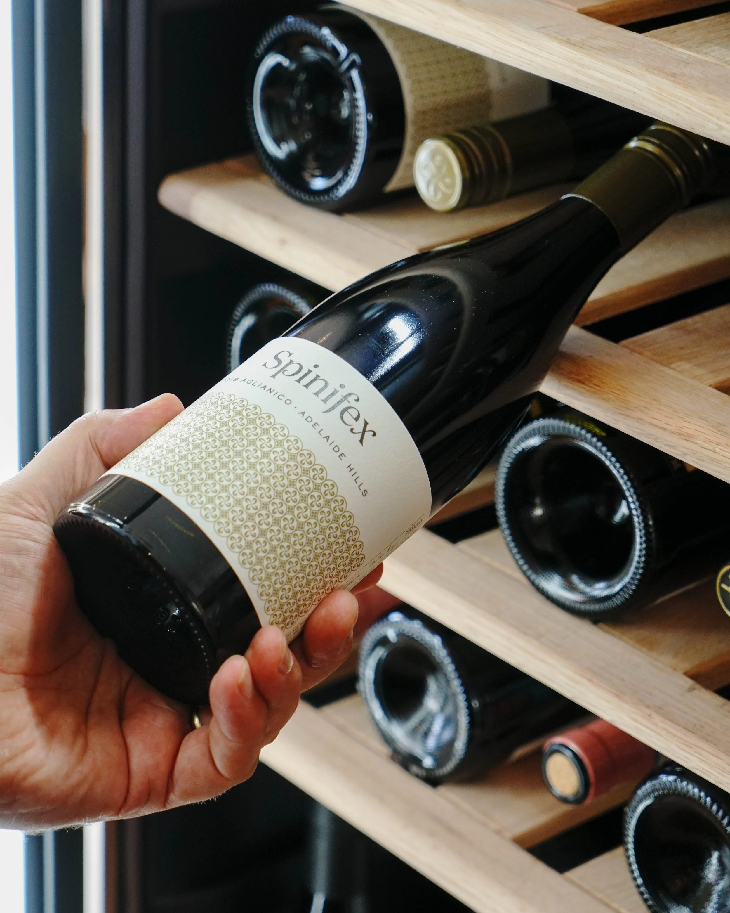 Person holding a bottle of Spinifex wine in front of a wine rack with other bottles.