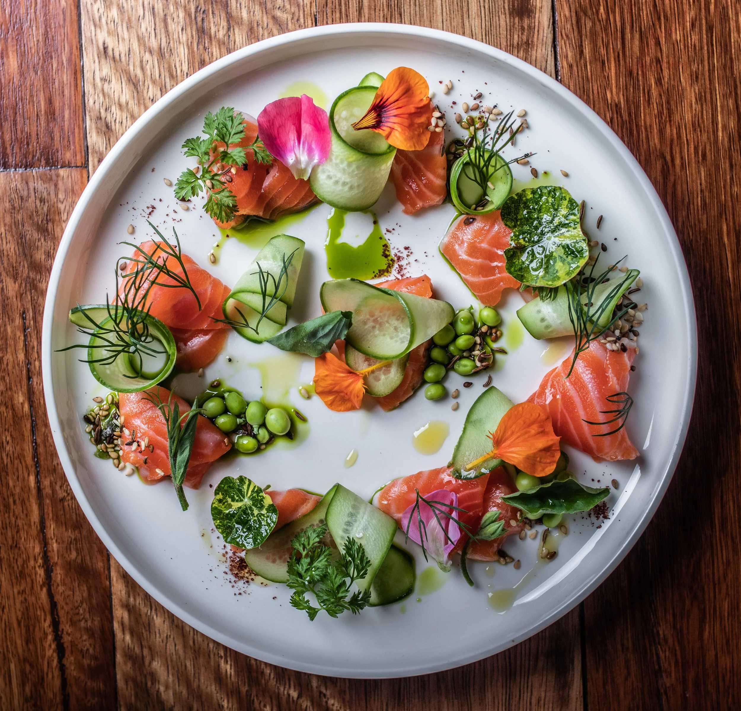 A white plate with thin slices of smoked salmon, cucumber ribbons, green peas, microgreens, edible flowers, and sesame seeds arranged as a gourmet appetizer.