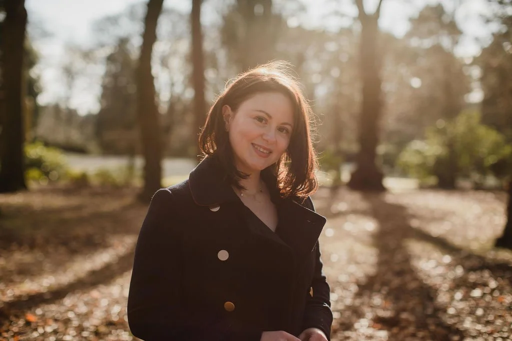 A woman with shoulder-length dark hair smiling outdoors in a wooded area during daylight, wearing a black jacket.