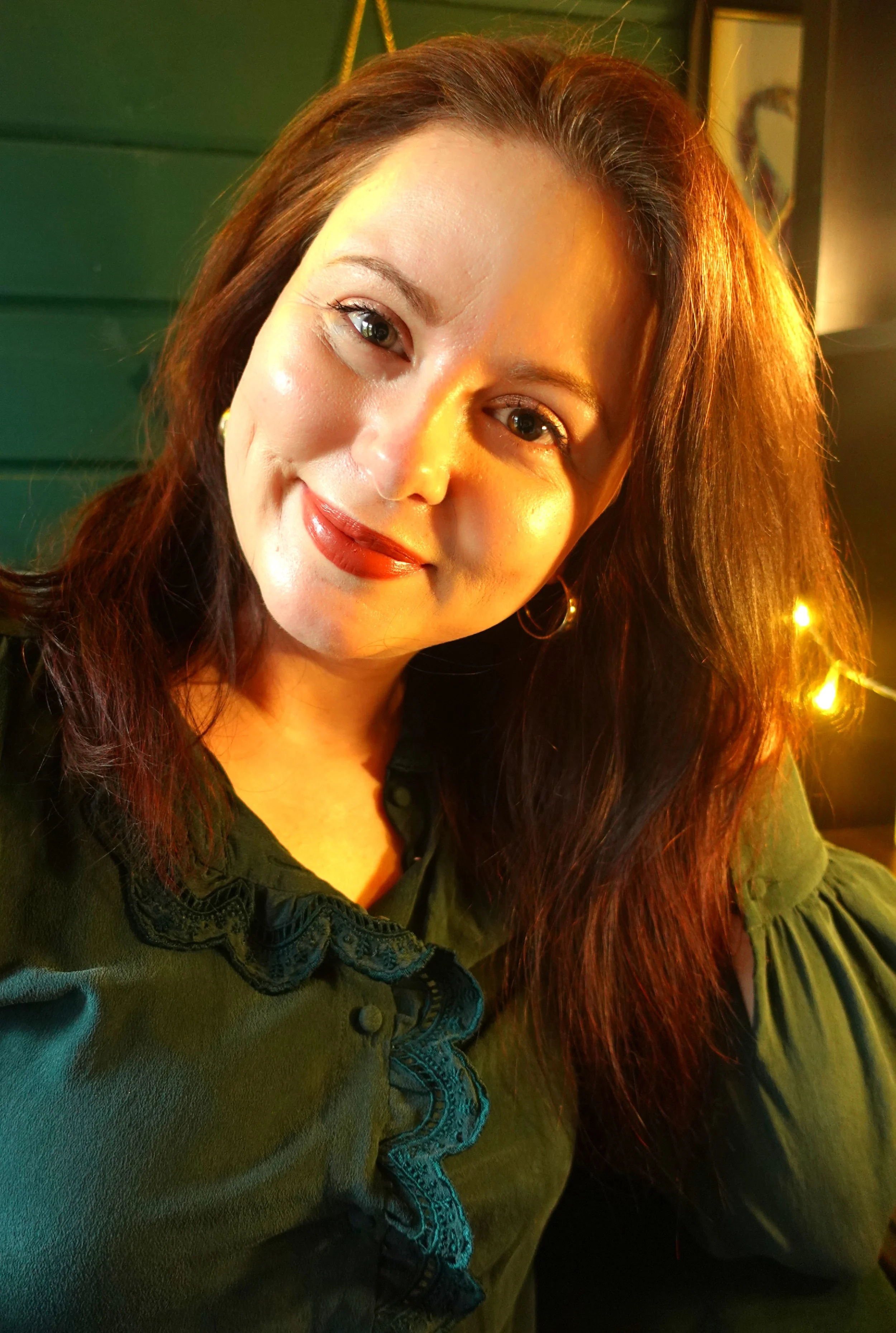 A woman with long, reddish-brown hair, wearing gold hoop earrings and a dark green blouse with lace trim, smiling softly in warm lighting.