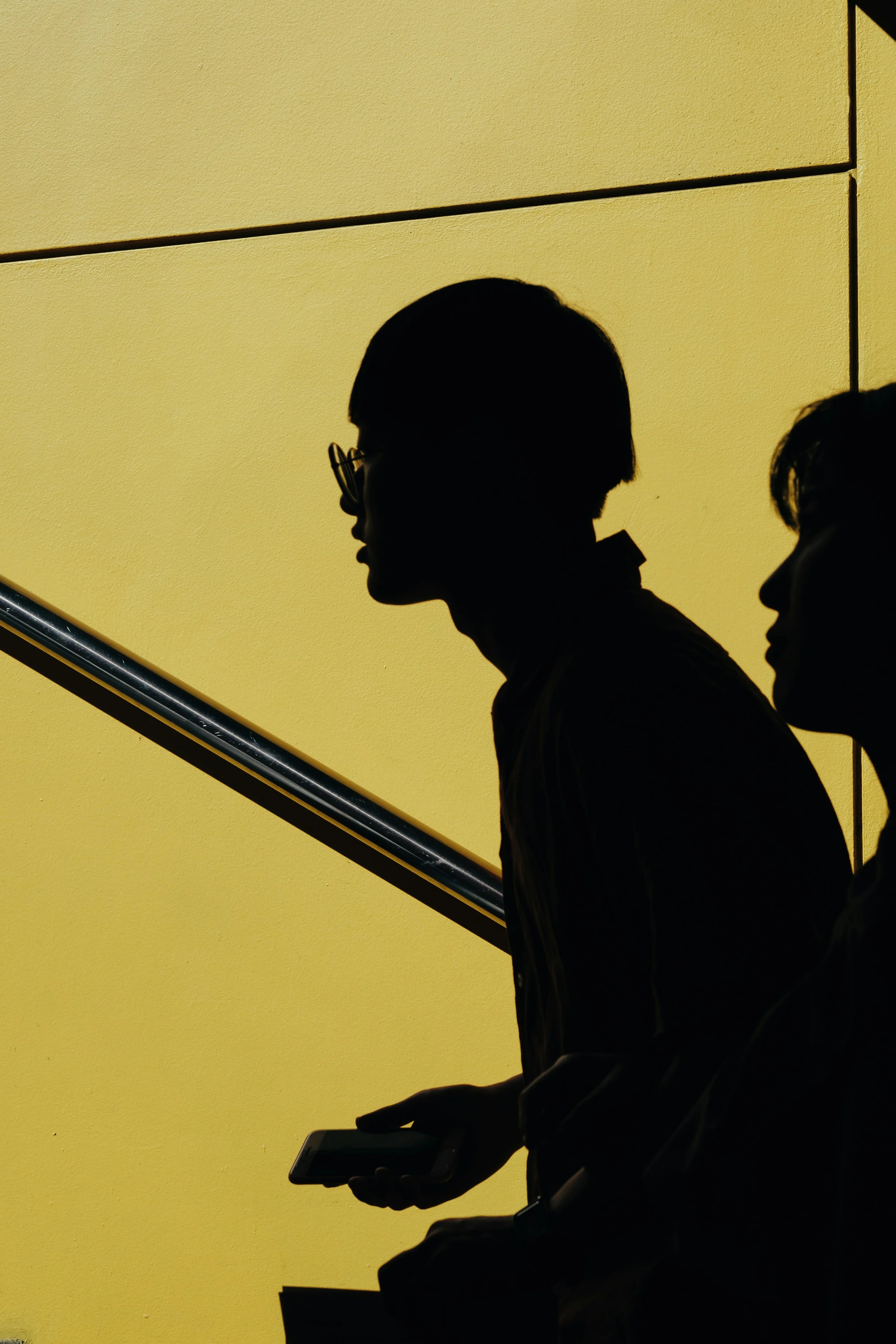 Silhouettes of two people walking down a staircase with a yellow wall in the background, one person holding a phone.