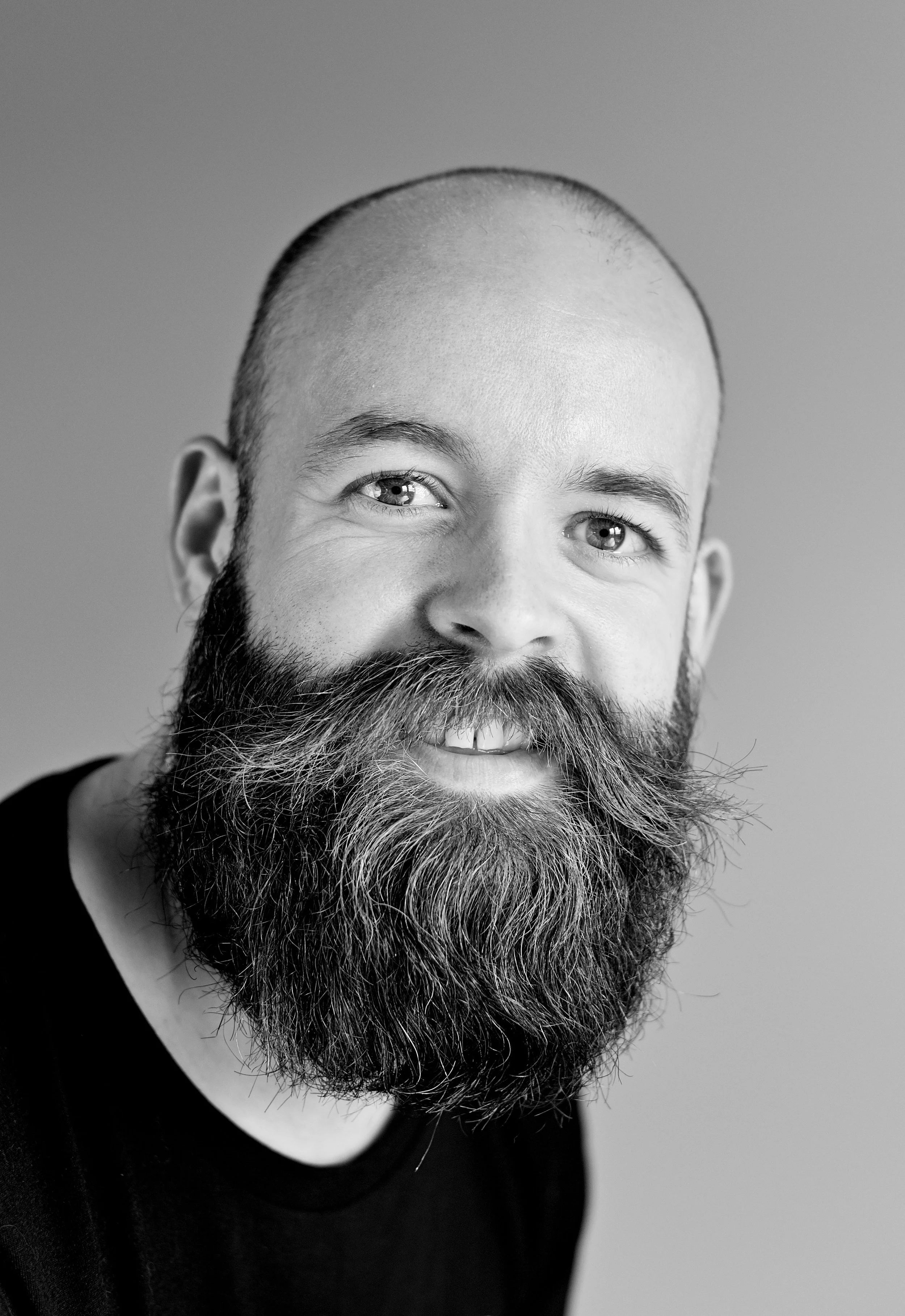 Close-up black and white portrait of a man with a shaved head, full beard, and smiling expression.