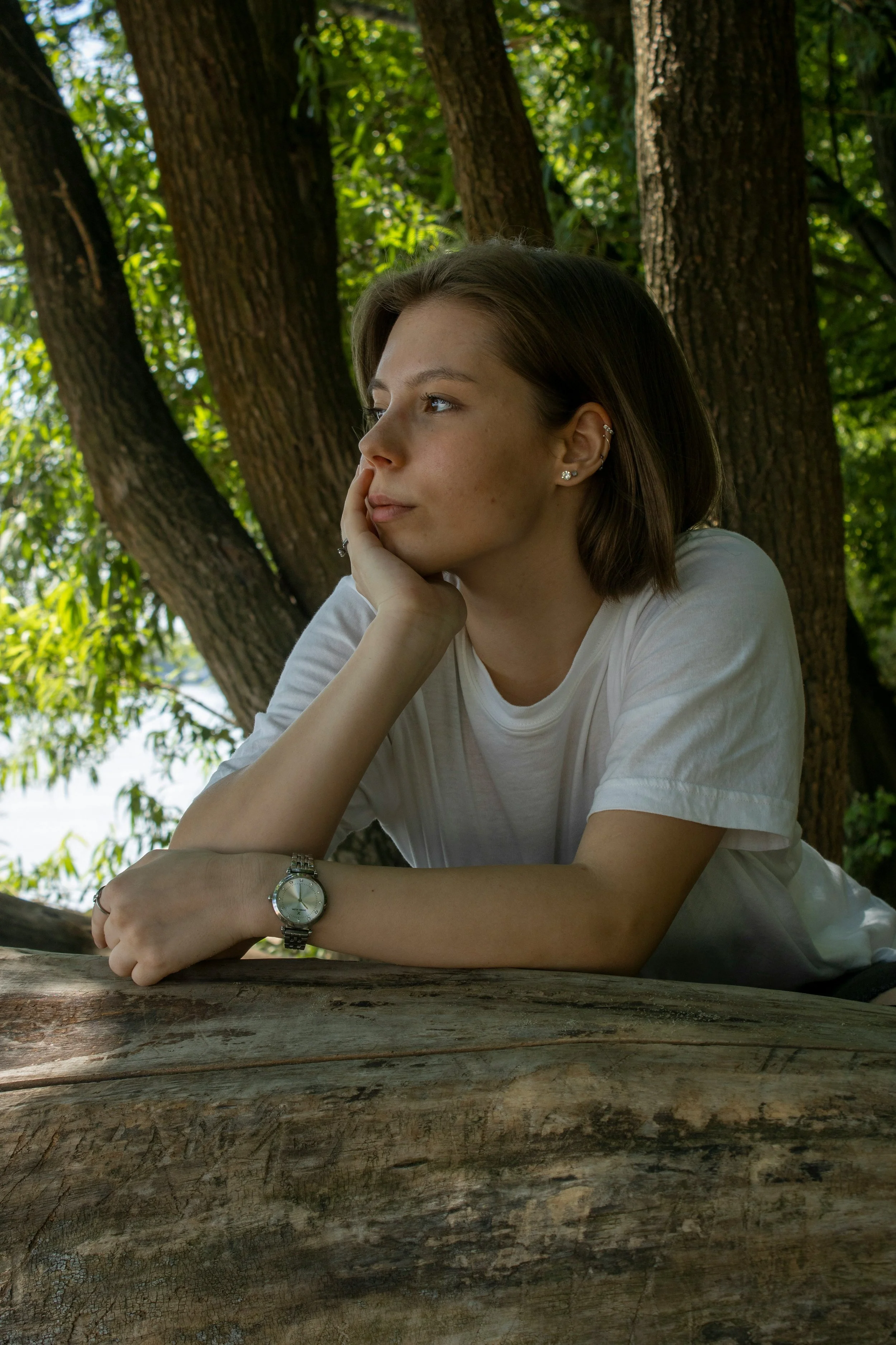 A young woman with short brown hair, wearing a white T-shirt and a silver watch, resting her chin on her hand while sitting or lying on a large log outdoors with green trees in the background.