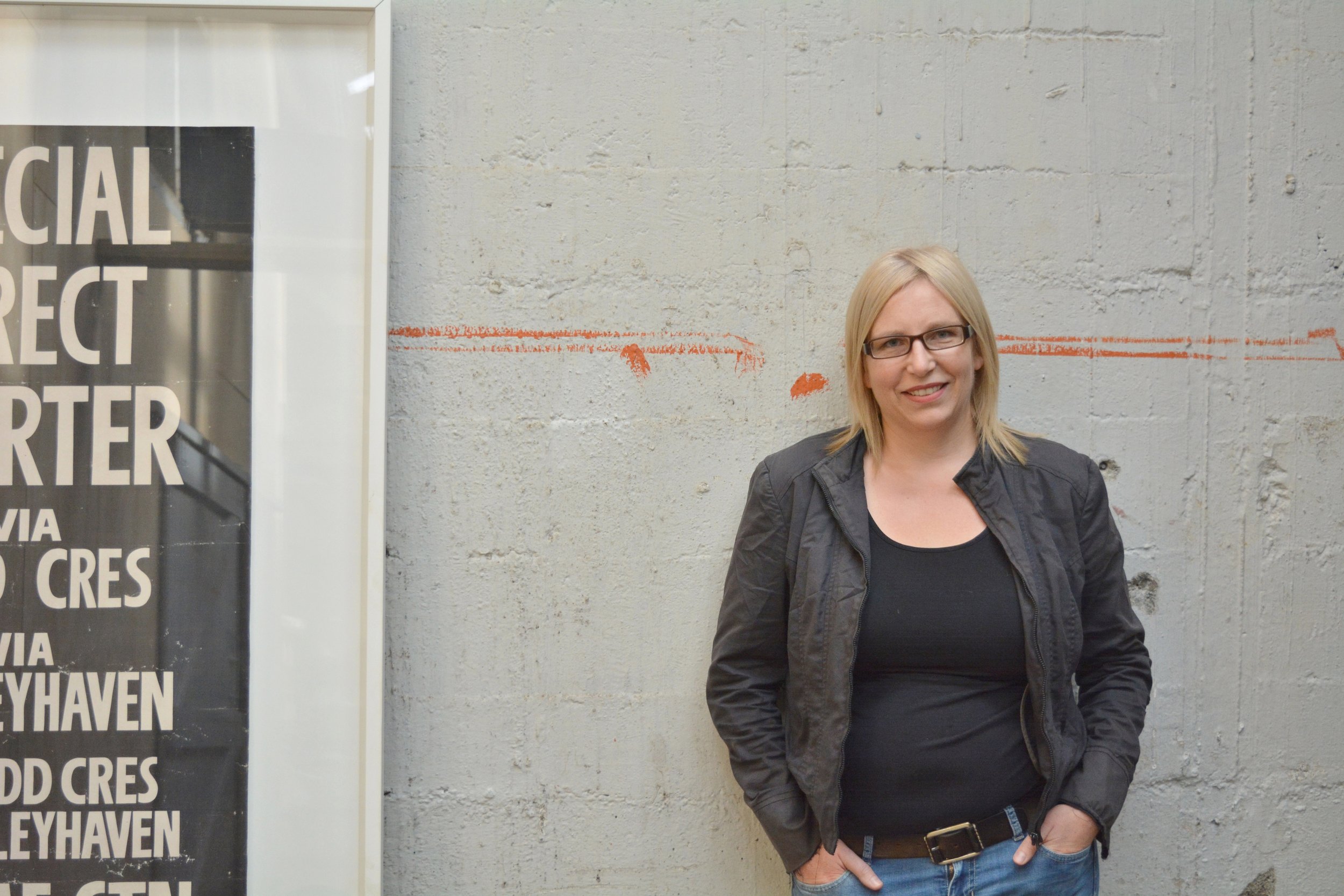 A woman with blonde hair, glasses, and a black jacket standing in front of a concrete wall with orange markings, smiling at the camera.