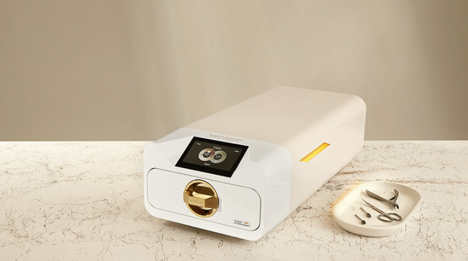 A white laboratory machine with a touchscreen display and a gold circular component sitting on a marble countertop. A small oval dish containing tweezers, scissors, and a small tool is positioned to the right of the machine.