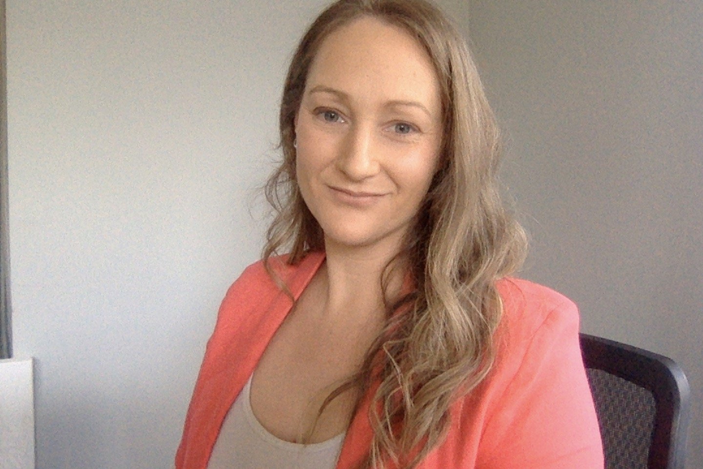 A woman with long, wavy blonde hair smiling at the camera, wearing a coral-colored blazer and a beige top, sitting in an office chair against a plain, light-colored wall.