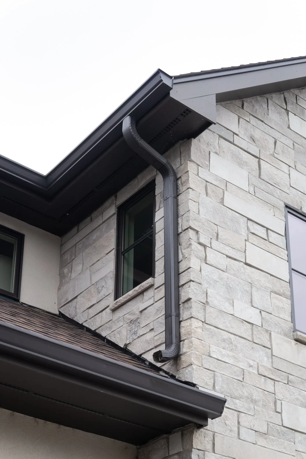 The exterior of a modern house with a stone facade, showing a black rain gutter, downspout, and section of roof, along with two windows. Roofing Company in Denver Colorado